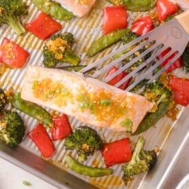 A metal spatula lifts a cooked salmon fillet garnished with green onions from a baking sheet with roasted broccoli, red bell peppers, and snap peas.