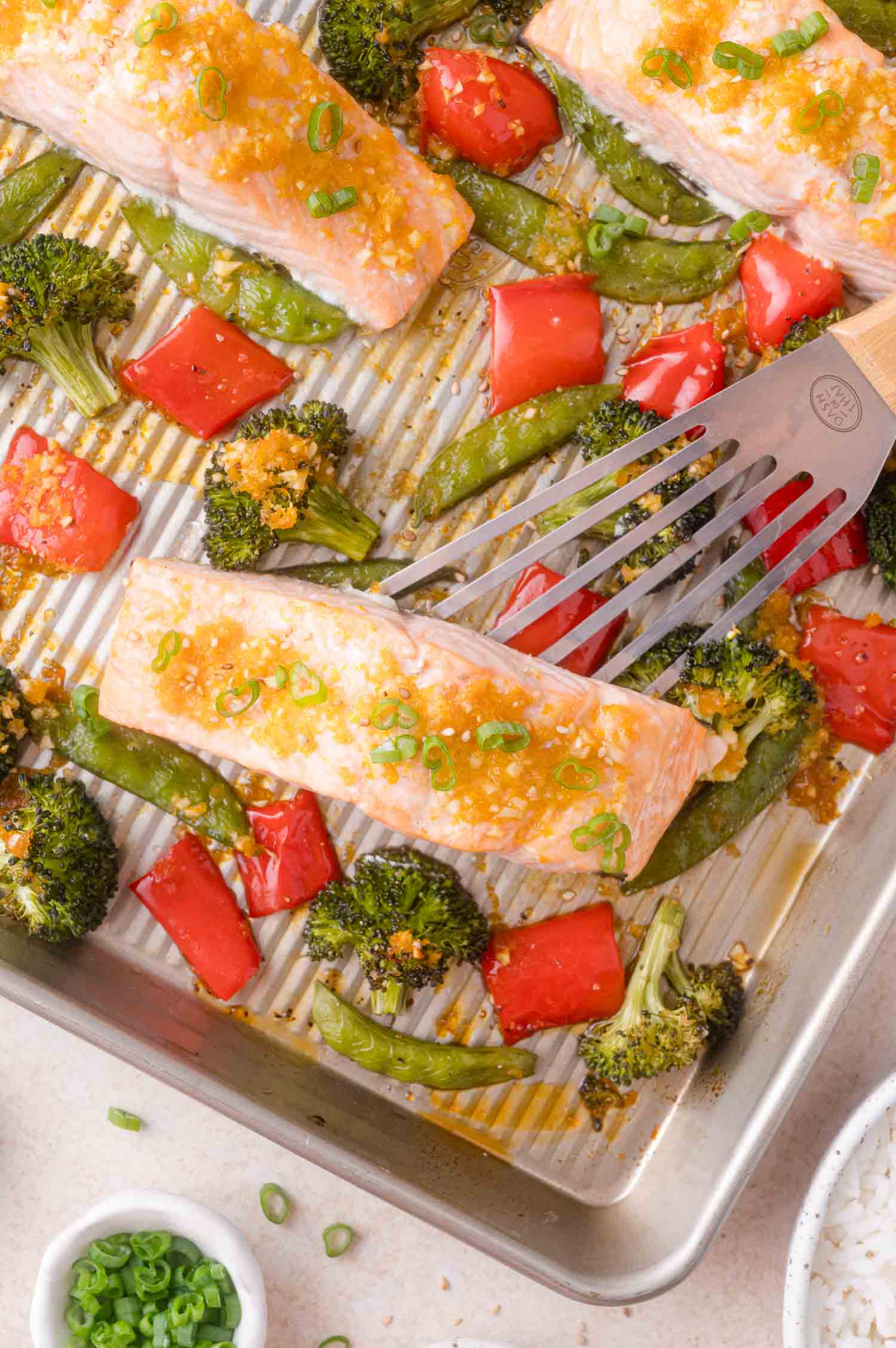 A metal spatula lifts a baked salmon fillet from a sheet pan with broccoli, red bell peppers, and snap peas.