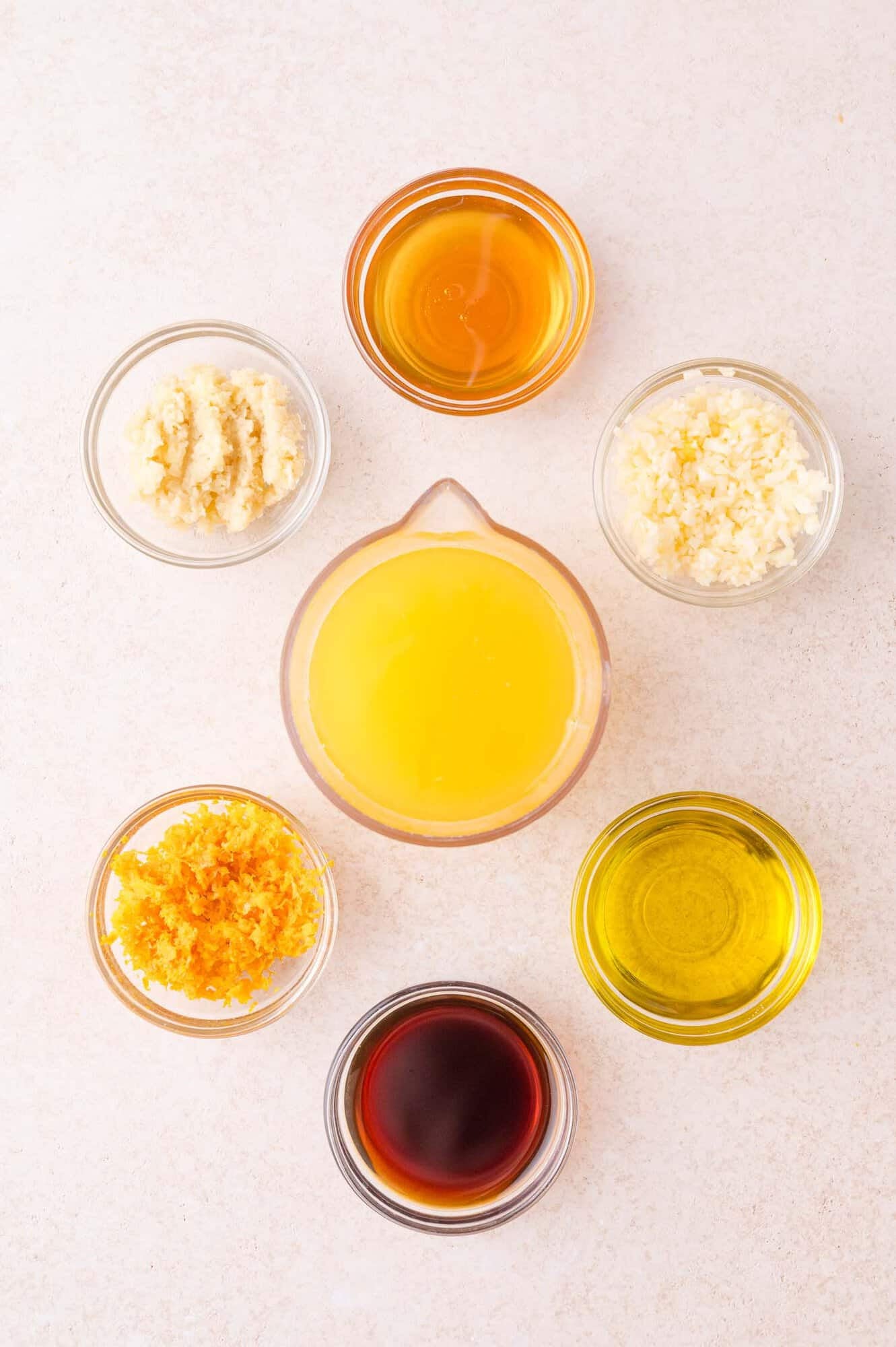 Seven small bowls containing various ingredients, including orange juice, olive oil, soy sauce, honey, grated ginger, grated garlic, and orange zest, arranged in a circle on a light surface.