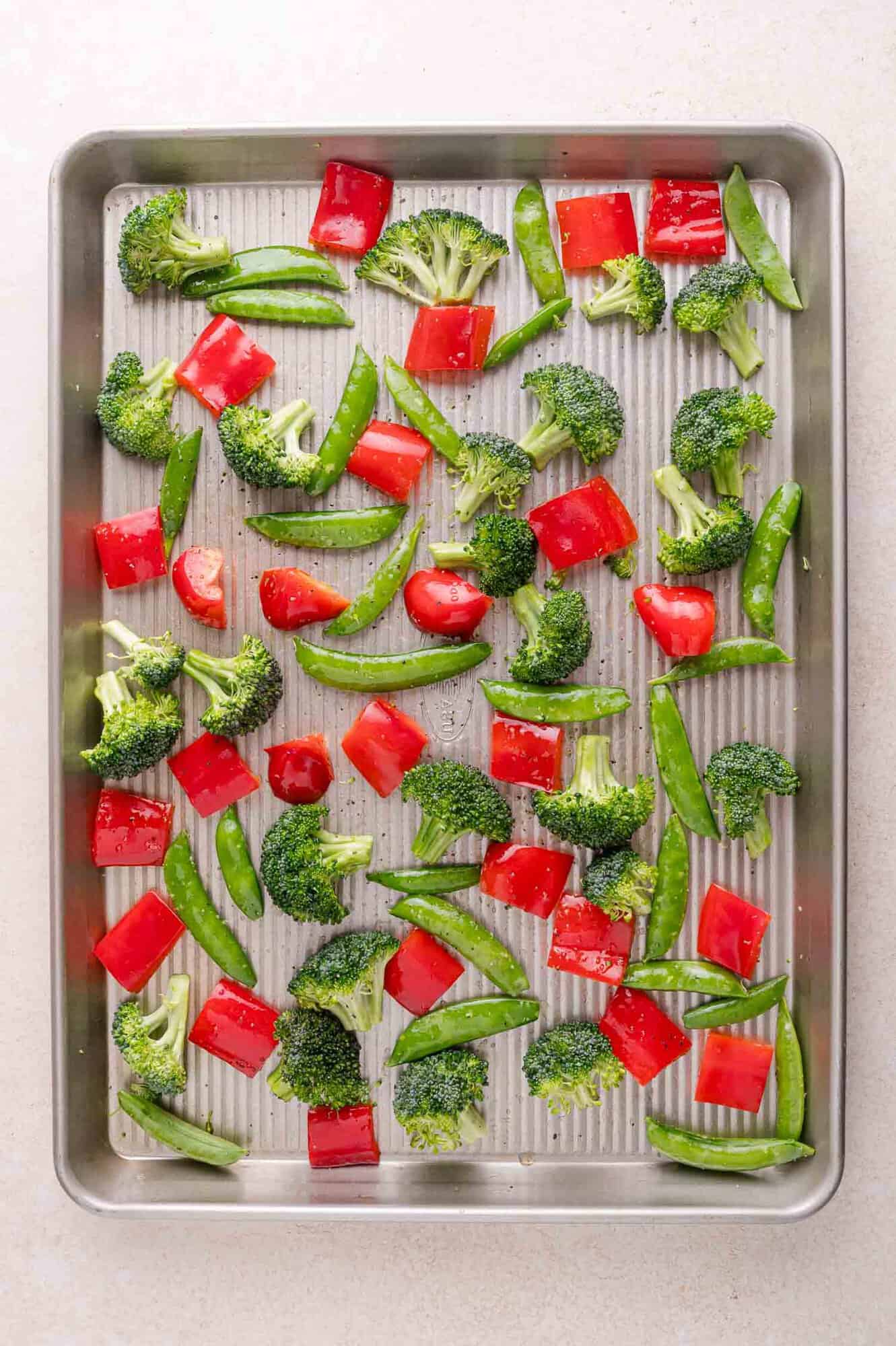 A baking sheet with raw broccoli florets, red bell pepper pieces, and snap peas spread evenly on a light-colored surface.