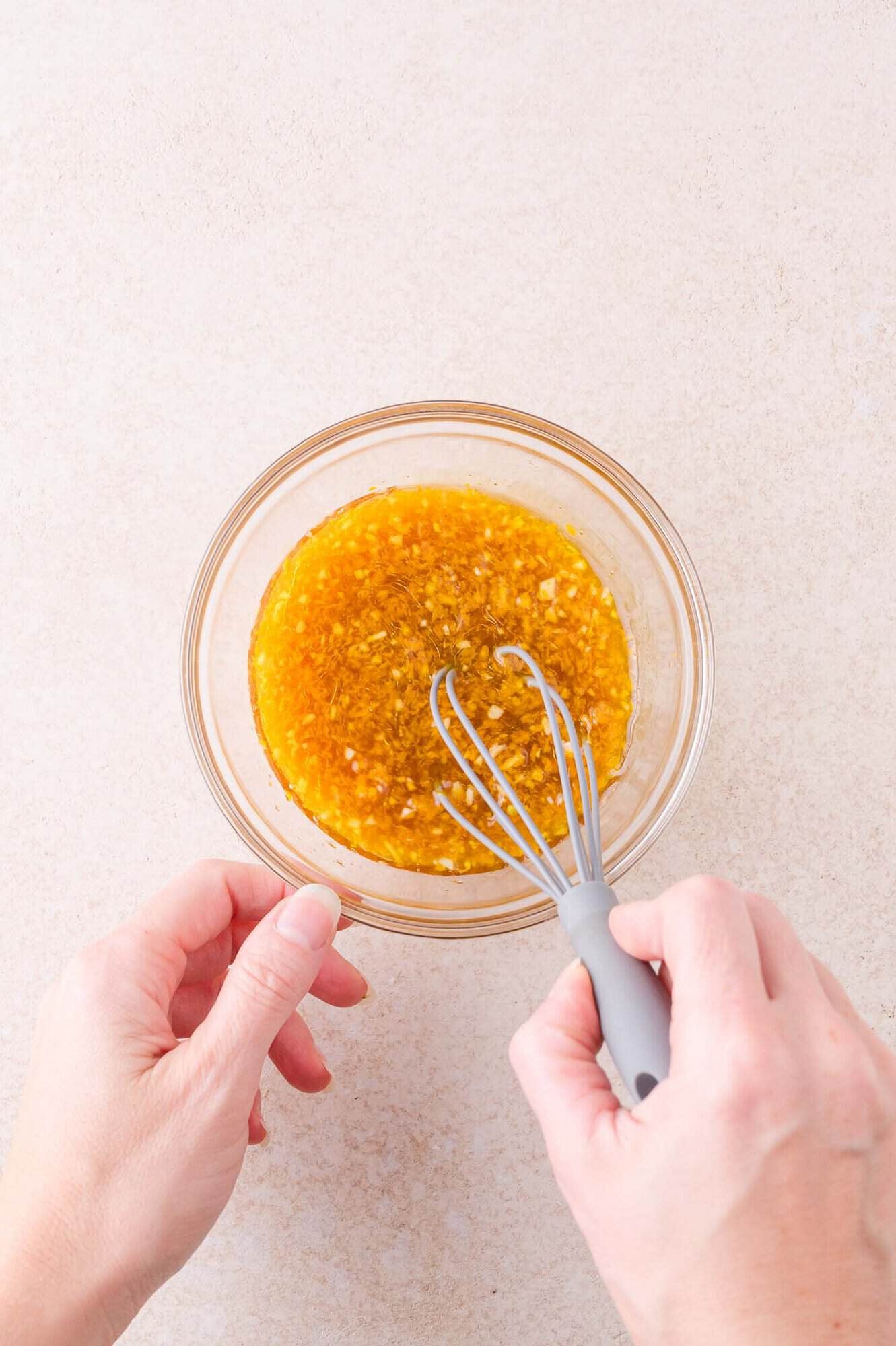 A person whisking a yellow-brown liquid mixture in a clear glass bowl on a light-colored surface.