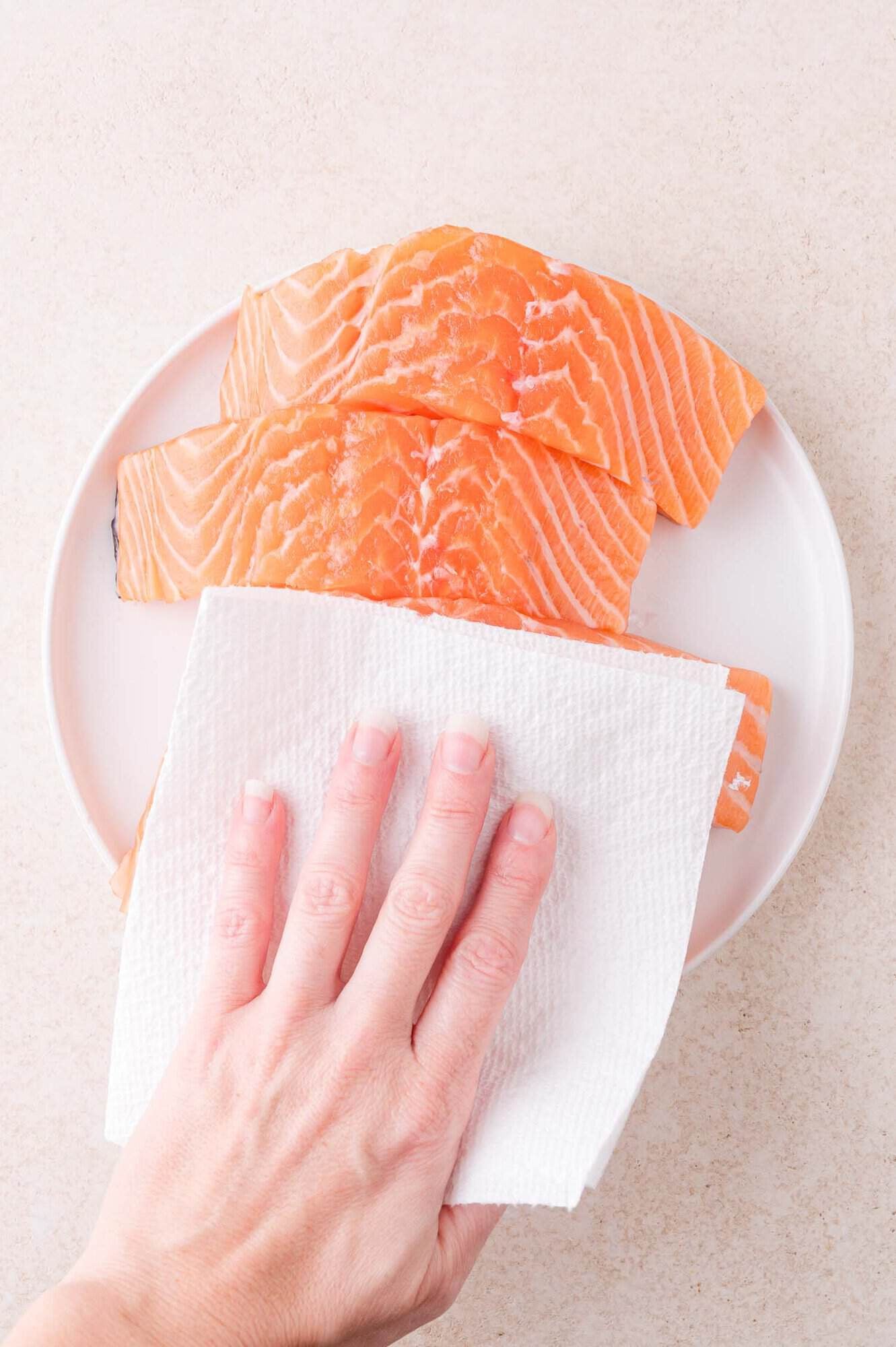 A hand uses a paper towel to pat dry raw salmon fillets on a white plate.
