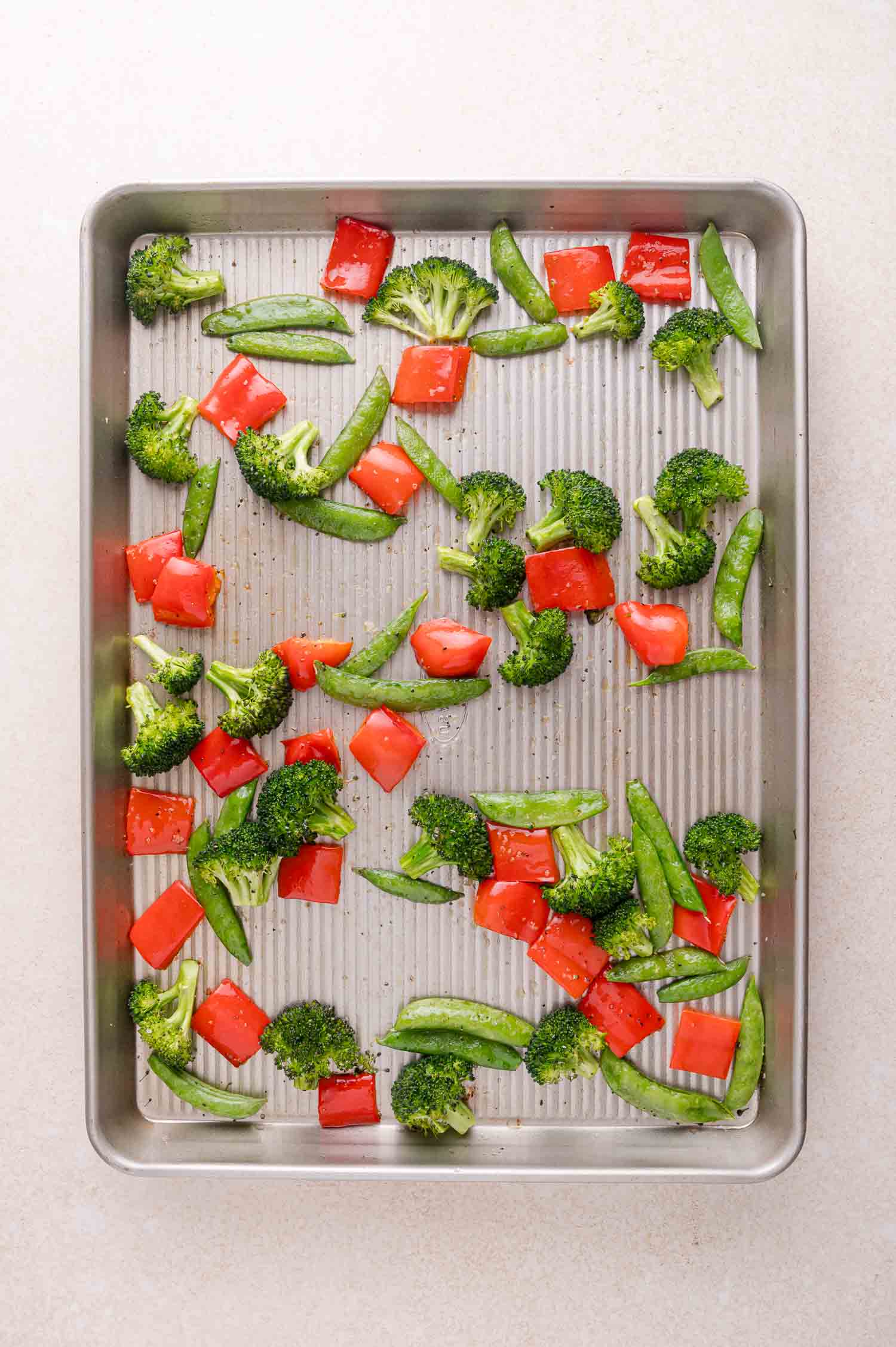 A baking sheet with raw broccoli florets, red bell pepper pieces, and snap peas spread evenly on the surface.
