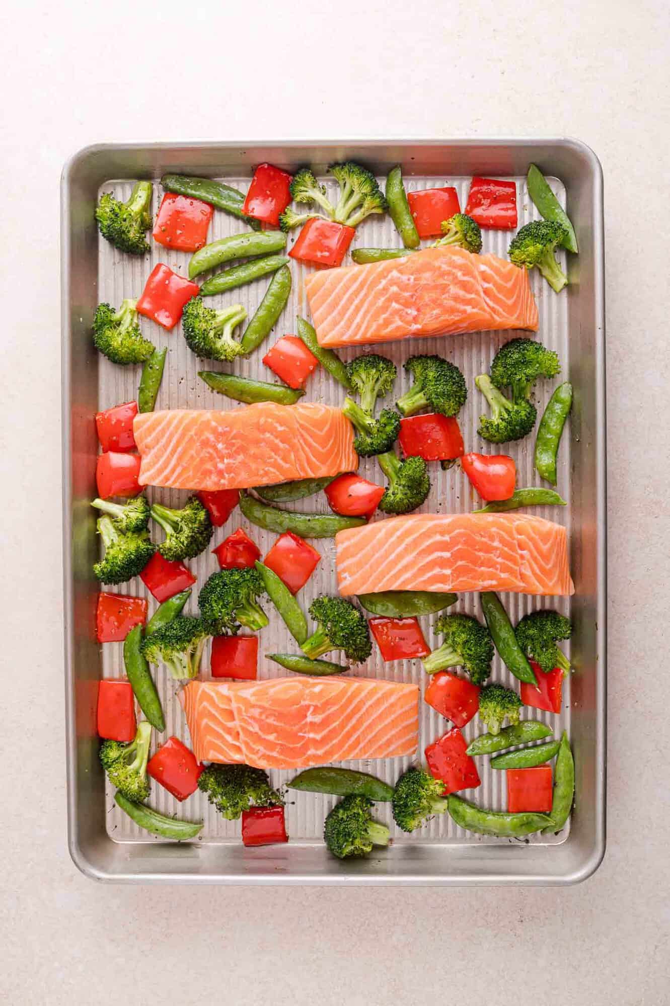 Four raw salmon fillets, broccoli, sugar snap peas, and red bell pepper pieces arranged on a metal baking sheet, ready for cooking.