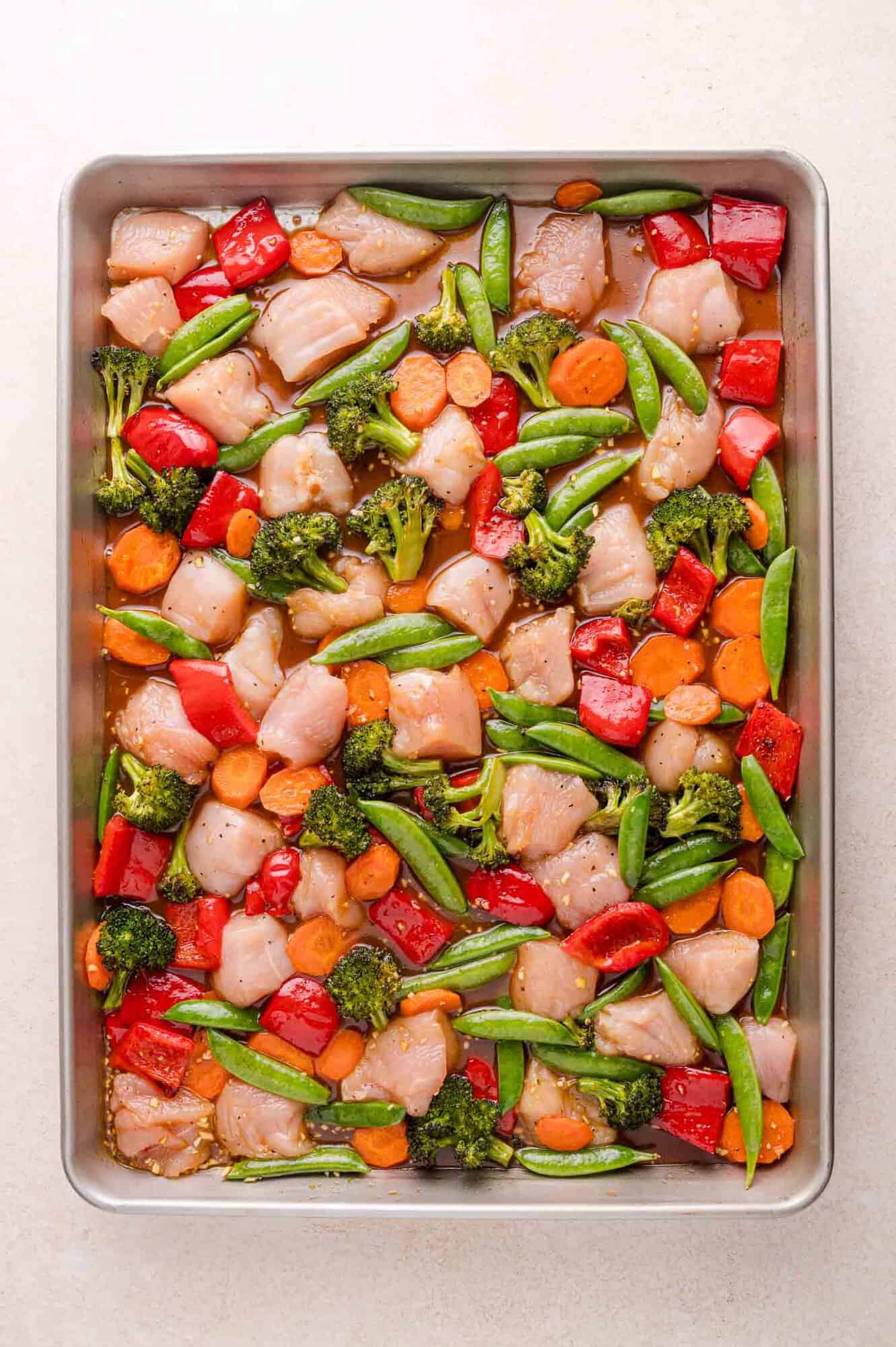 A sheet pan filled with raw diced chicken, broccoli, red bell peppers, snap peas, and carrot slices, ready to be cooked.