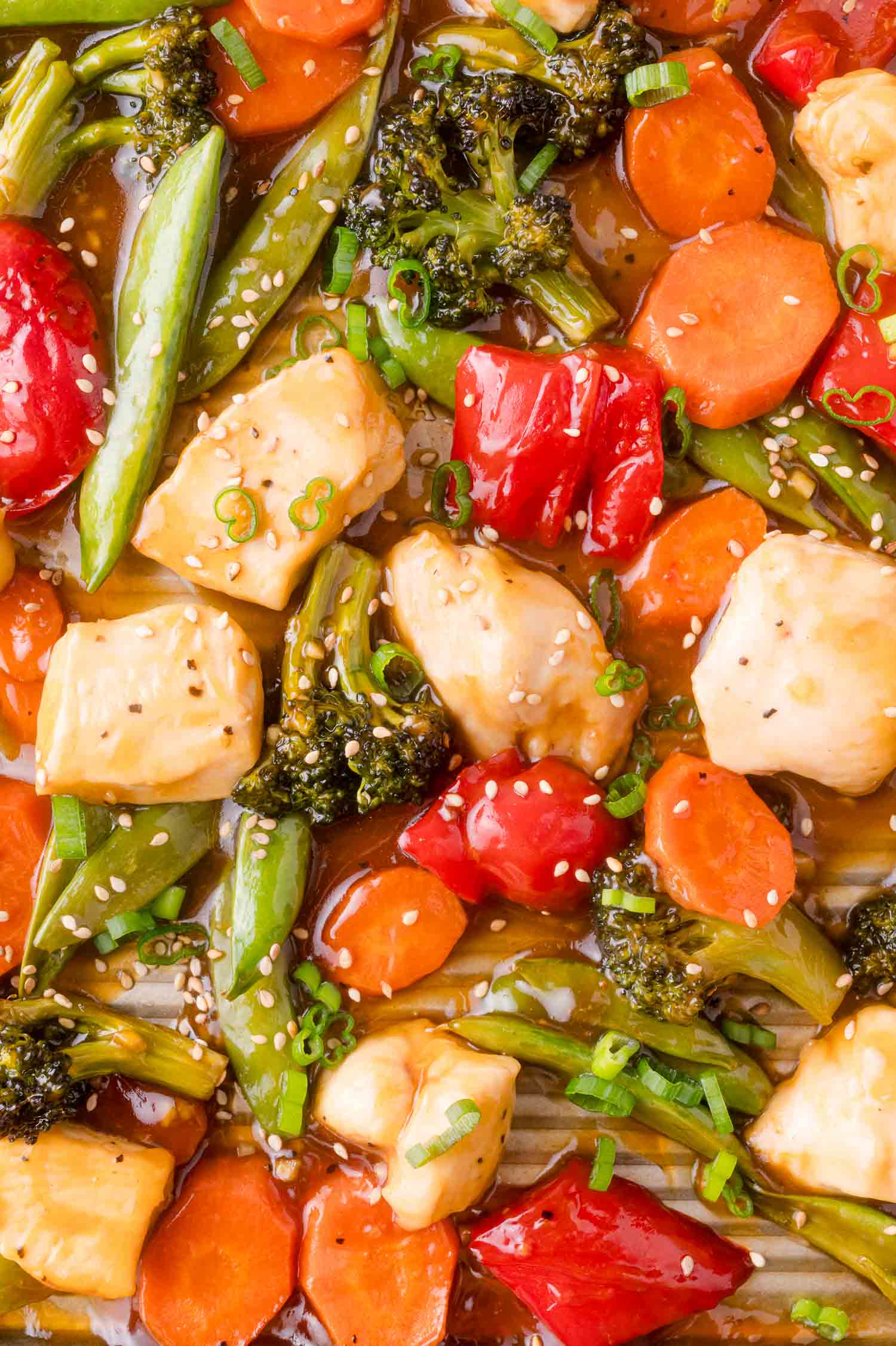 Close-up of a colorful stir-fry with chicken, broccoli, carrots, red bell pepper, snap peas, and cherry tomatoes in sauce, garnished with sesame seeds and chopped green onions.
