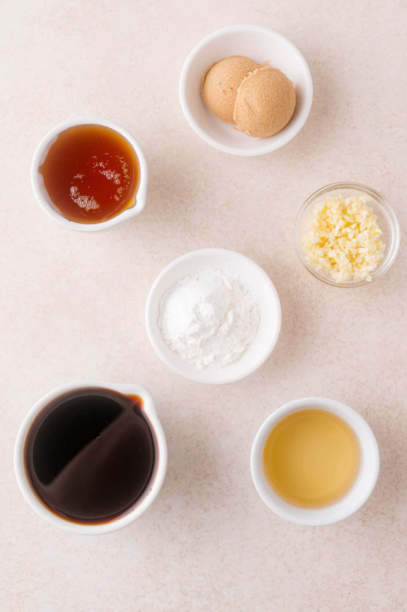 Six small bowls containing soy sauce, brown sugar, minced garlic, honey, cornstarch, and rice vinegar arranged on a light surface.