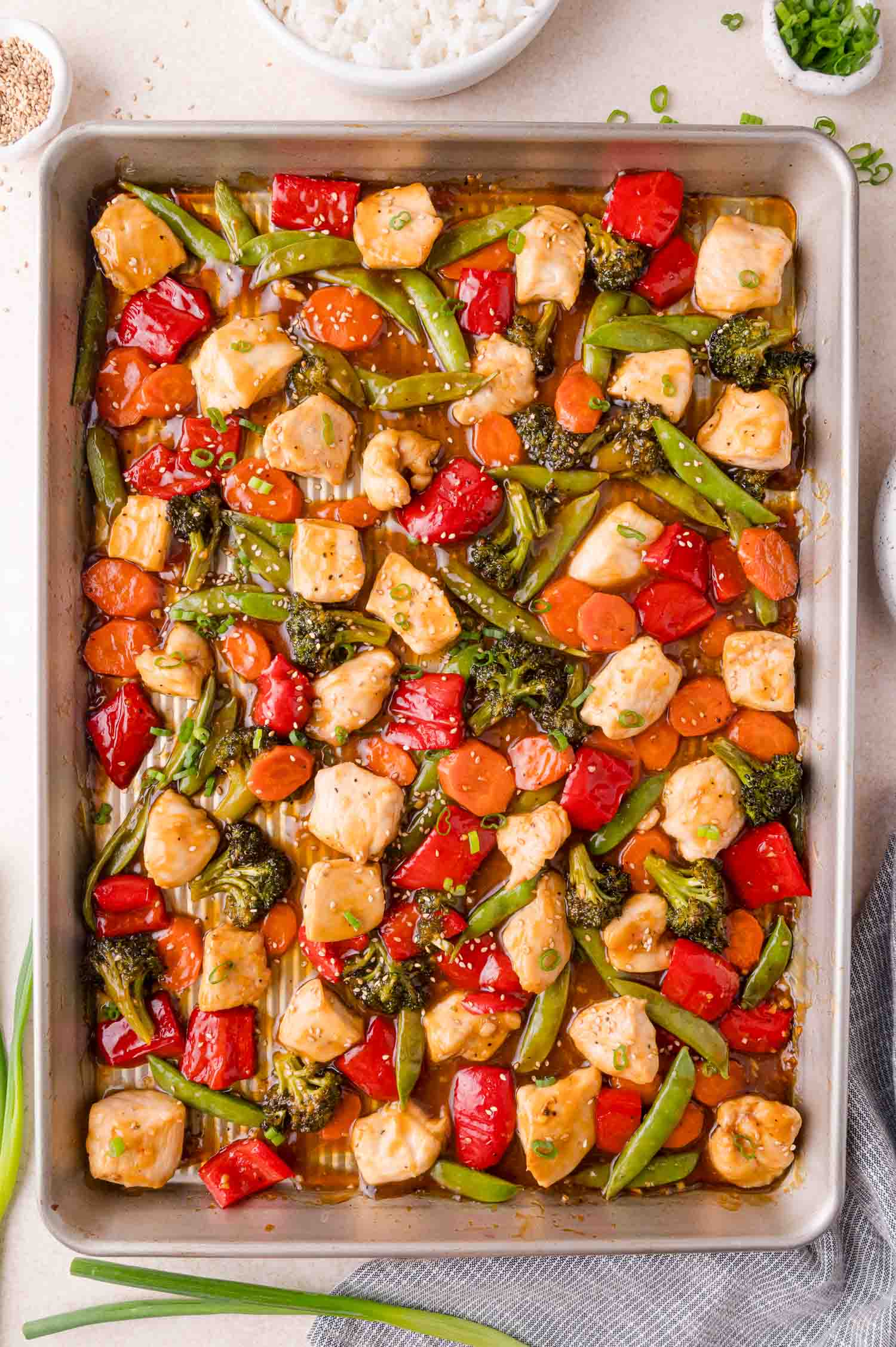 A sheet pan filled with cooked chicken pieces, broccoli, red bell peppers, snap peas, and carrots in a savory sauce. Bowls of rice and green onions are nearby.
