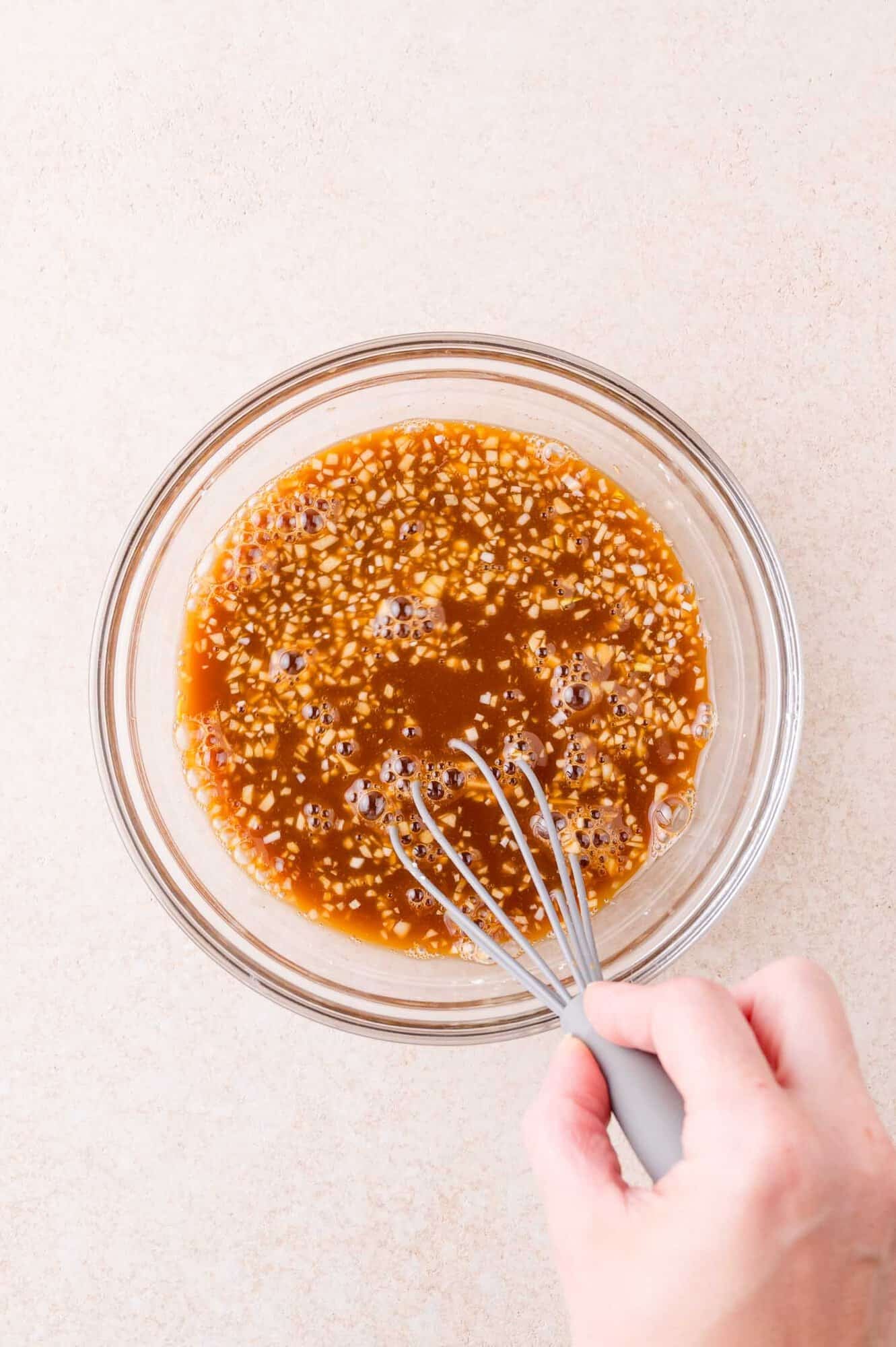 A hand holding a whisk mixes a brown sauce with visible garlic in a clear glass bowl on a light-colored surface.