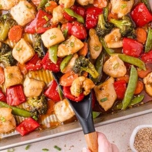 A tray of chicken stir-fry with broccoli, bell peppers, snap peas, carrots, and sauce, being served with a black spatula.