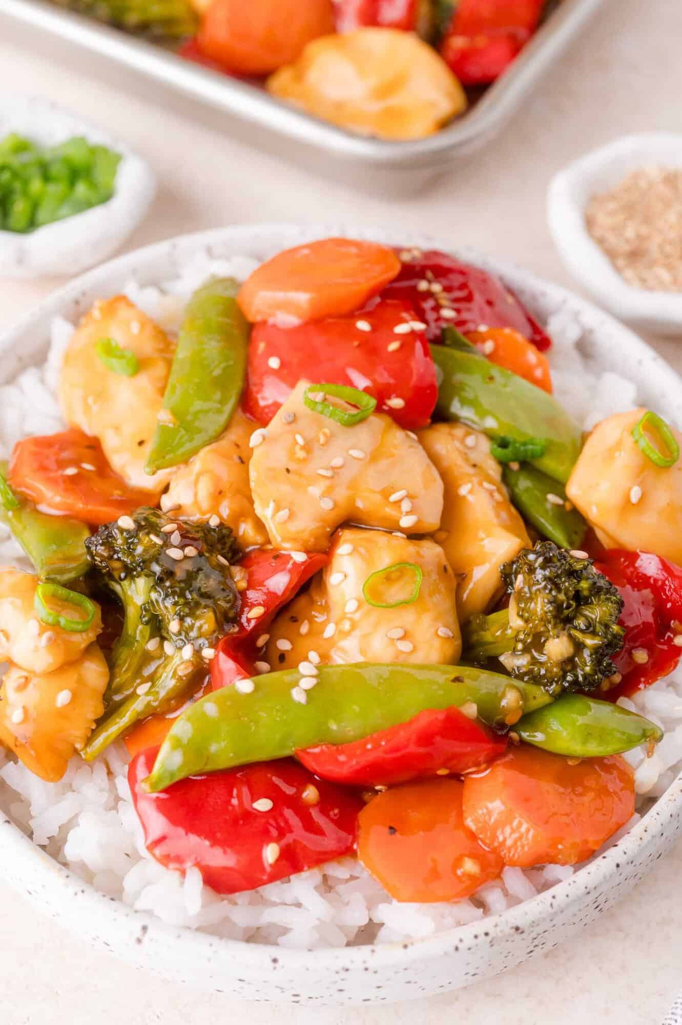 A bowl of white rice topped with stir-fried chicken, broccoli, snap peas, red bell peppers, carrots, and sesame seeds.