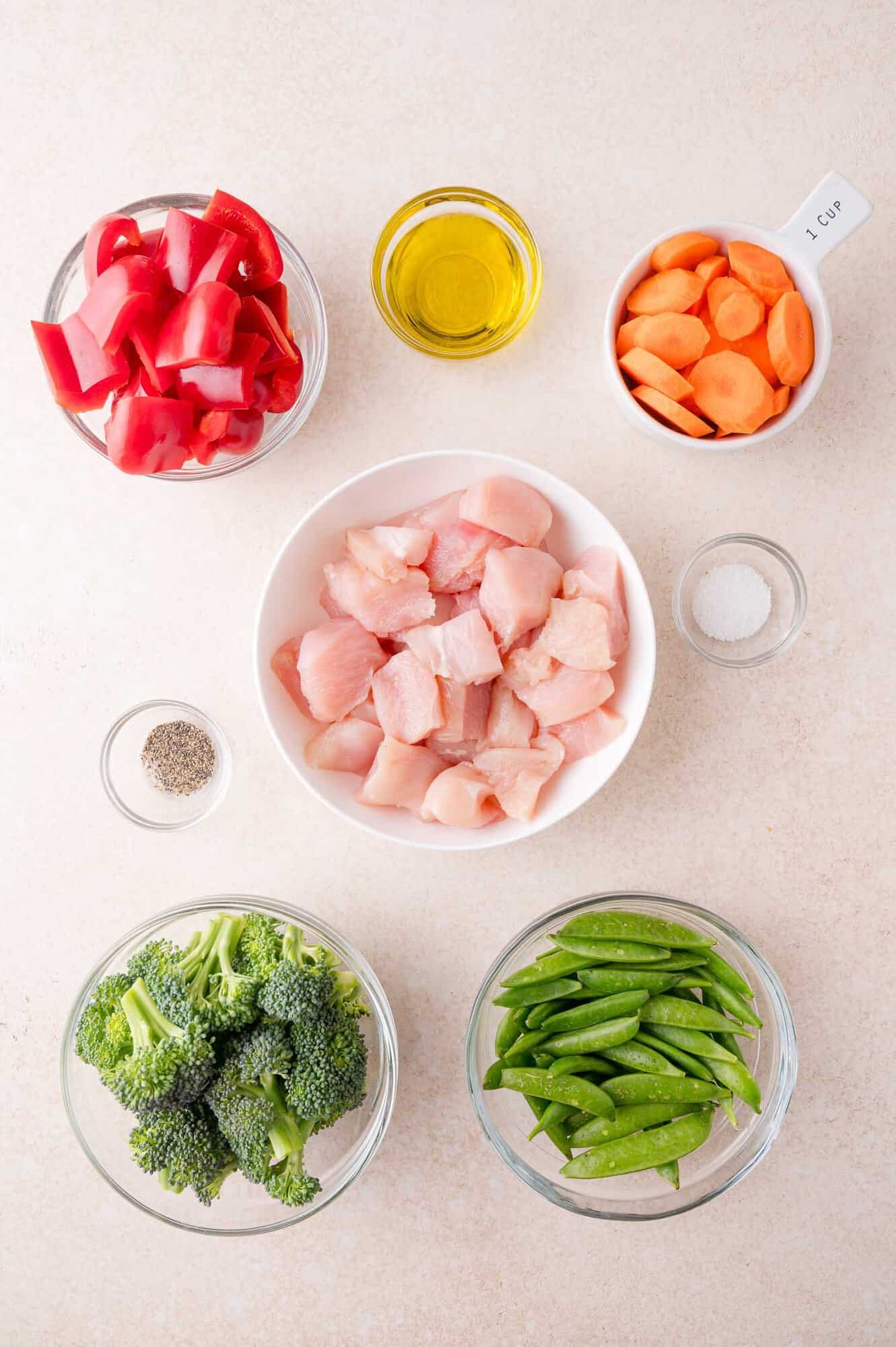 Raw diced chicken, broccoli, snap peas, red bell peppers, sliced carrots, olive oil, salt, and pepper arranged in separate bowls on a light surface.