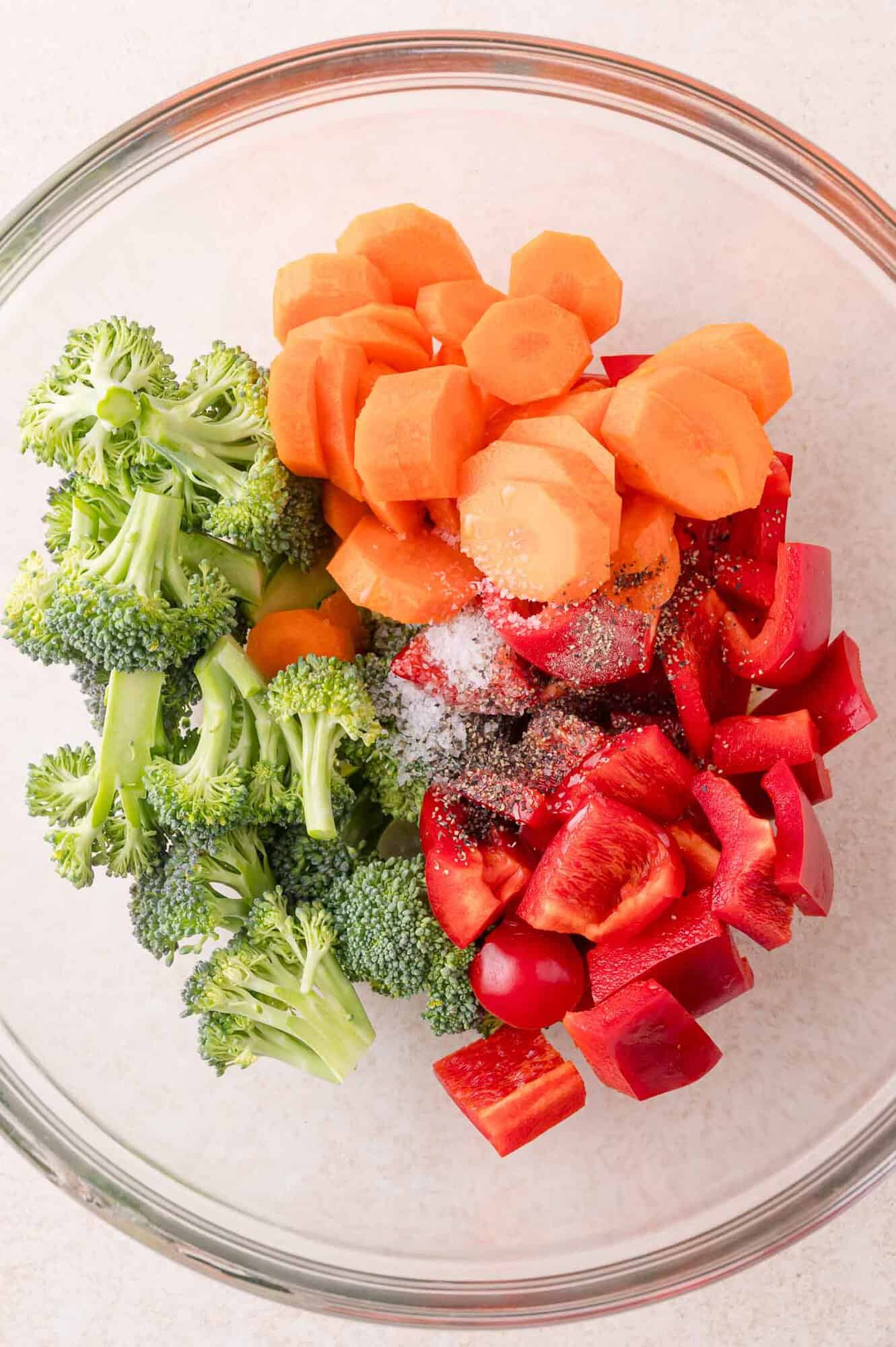 A glass bowl containing chopped broccoli, sliced carrots, and diced red bell pepper, topped with salt and pepper.