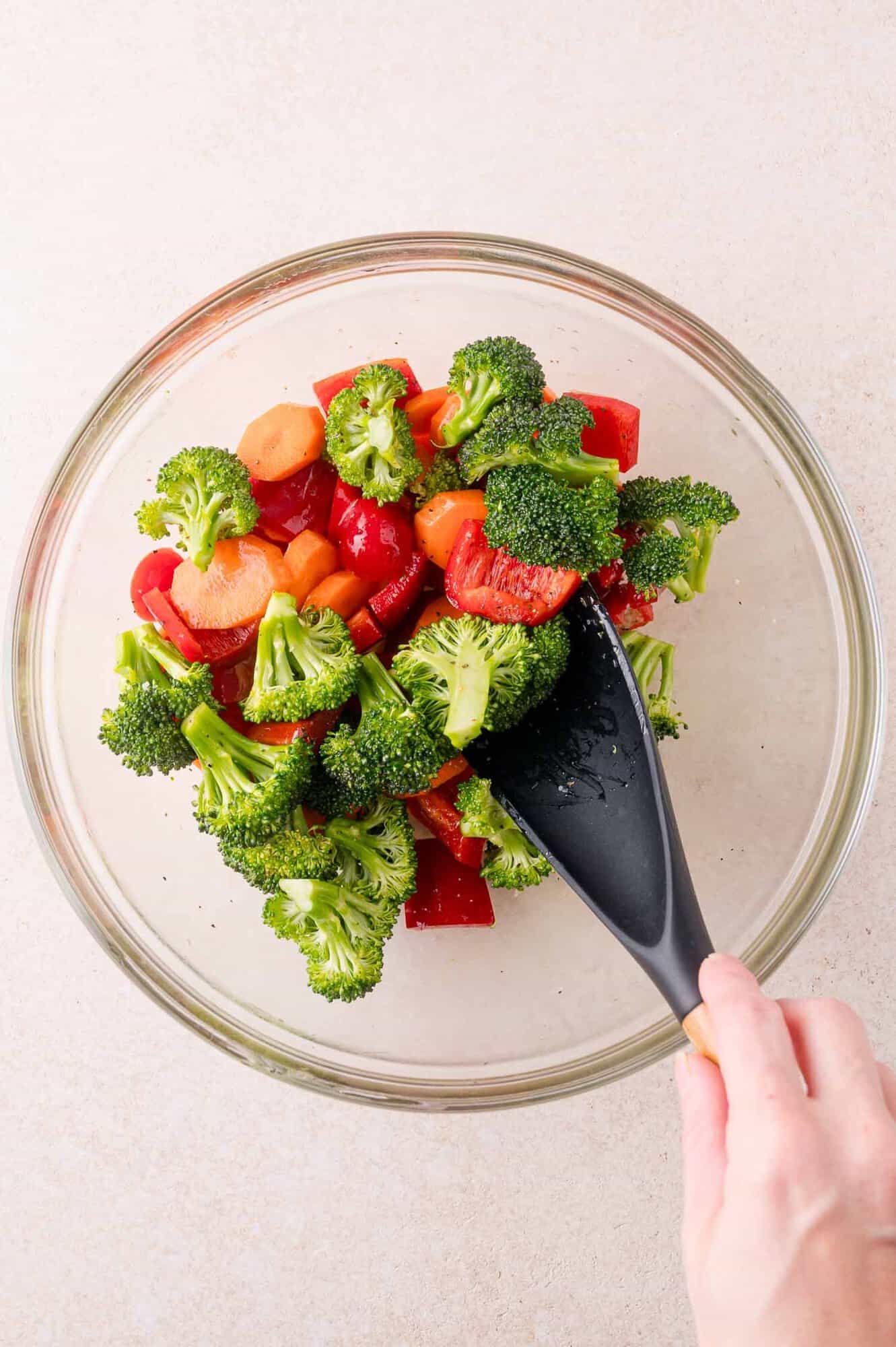 A hand uses a black spatula to mix fresh broccoli, red bell pepper, and sliced carrots in a glass bowl.