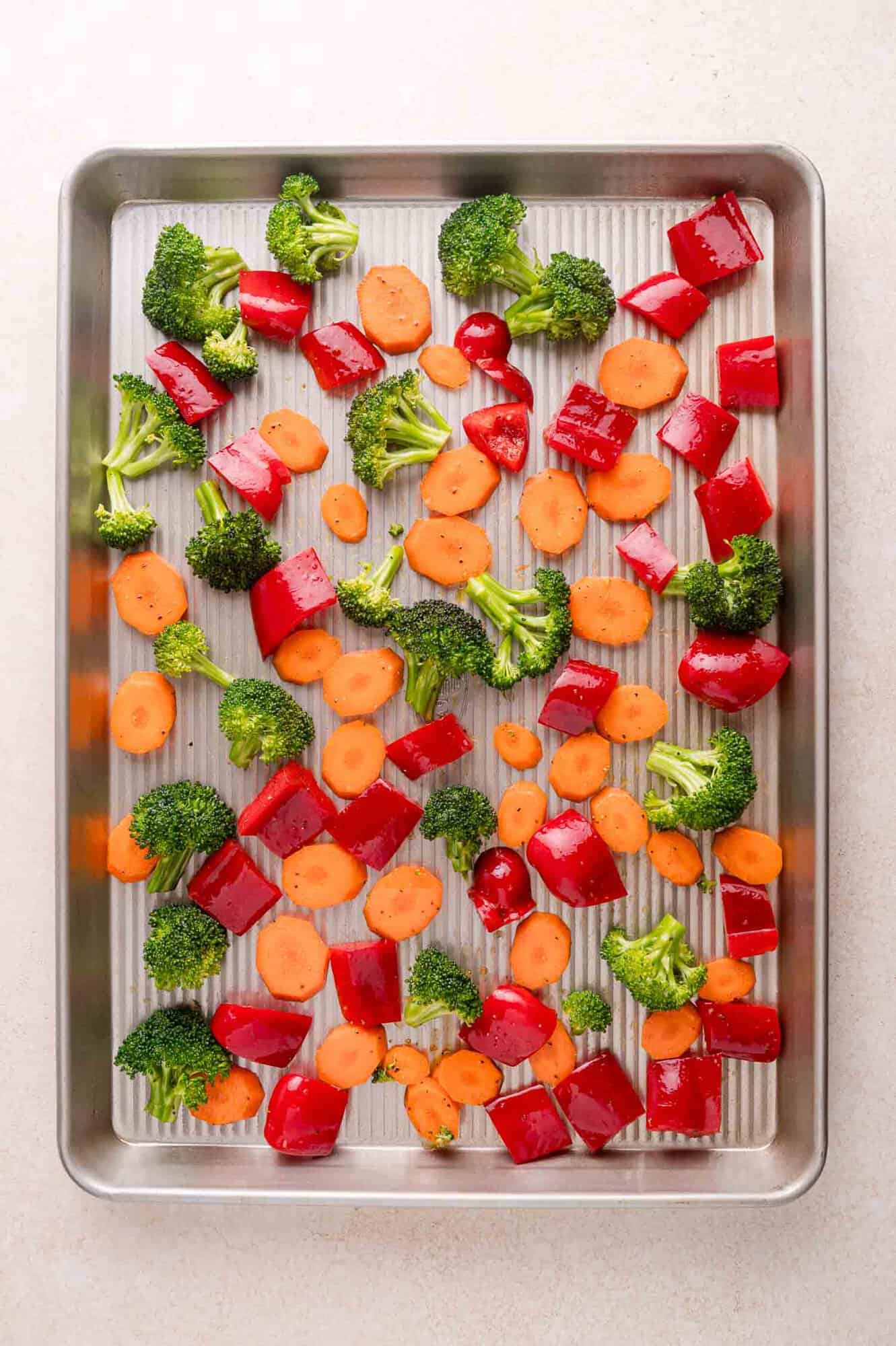 Chopped broccoli, red bell pepper, and carrot pieces are spread evenly on a metal baking sheet, ready for roasting.
