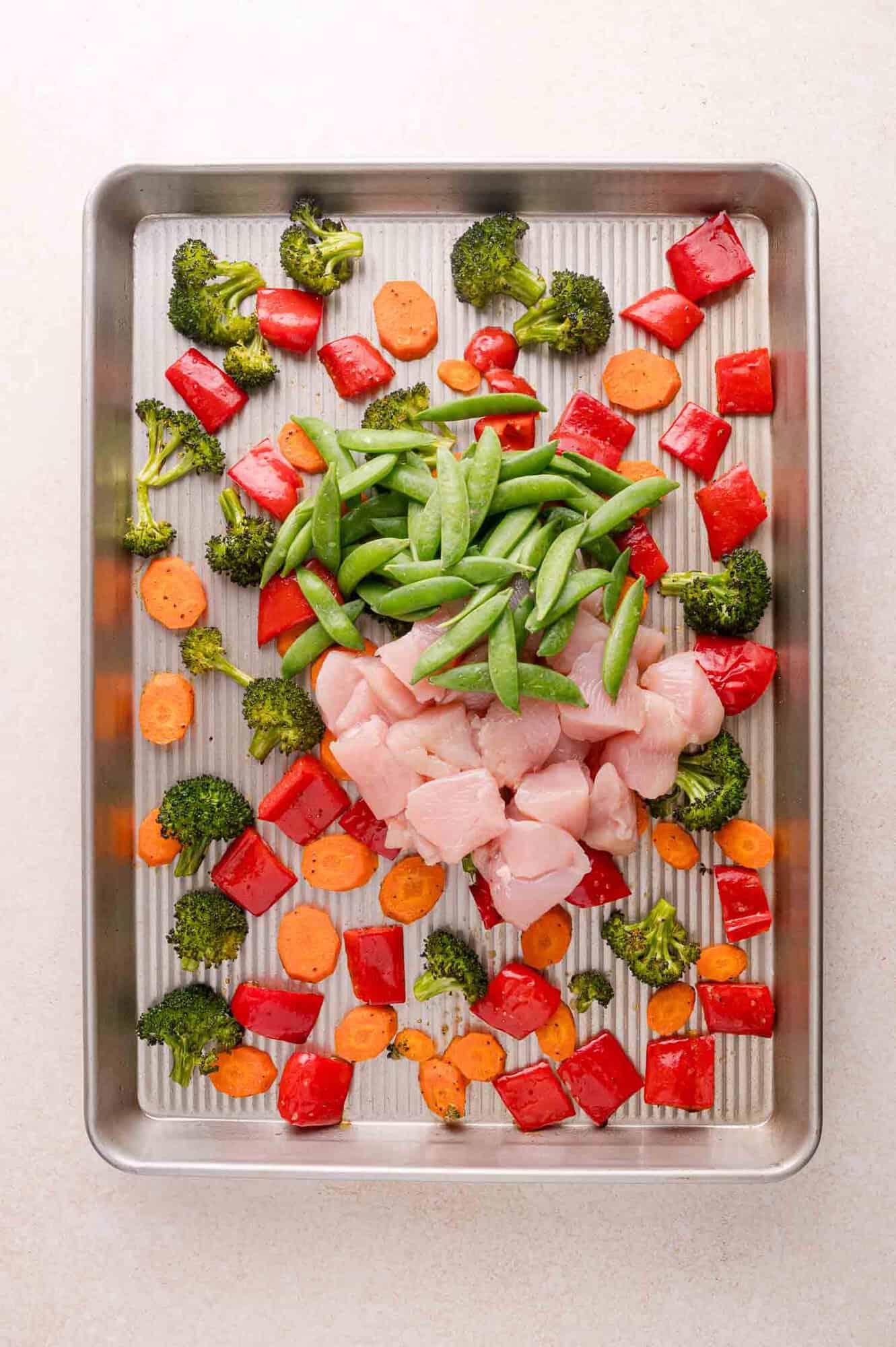 Raw chicken pieces, sugar snap peas, chopped red bell pepper, broccoli florets, and sliced carrots arranged on a metal baking sheet.