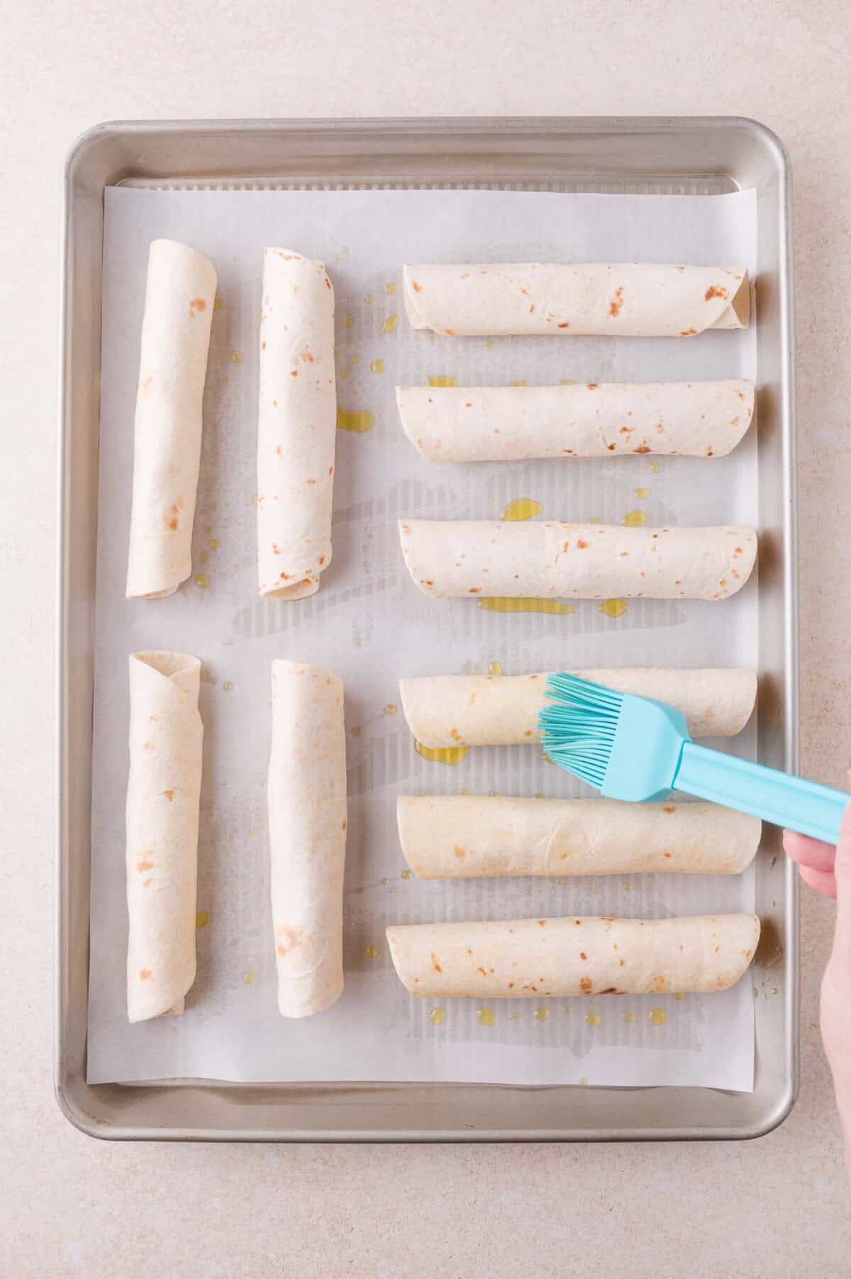 A hand brushes oil onto rolled tortillas arranged on a parchment-lined baking sheet.