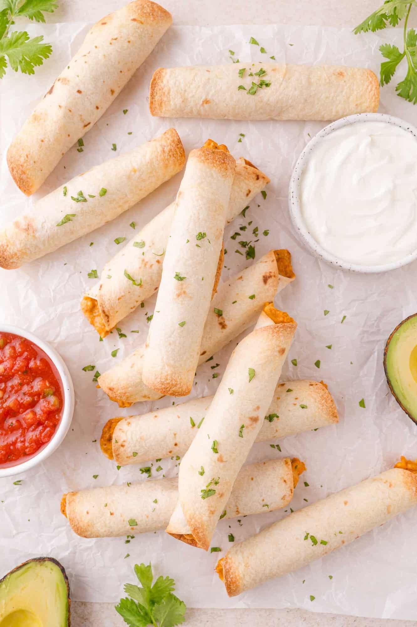 A pile of baked taquitos on parchment paper, garnished with chopped herbs, served with bowls of salsa and sour cream, and avocado halves.