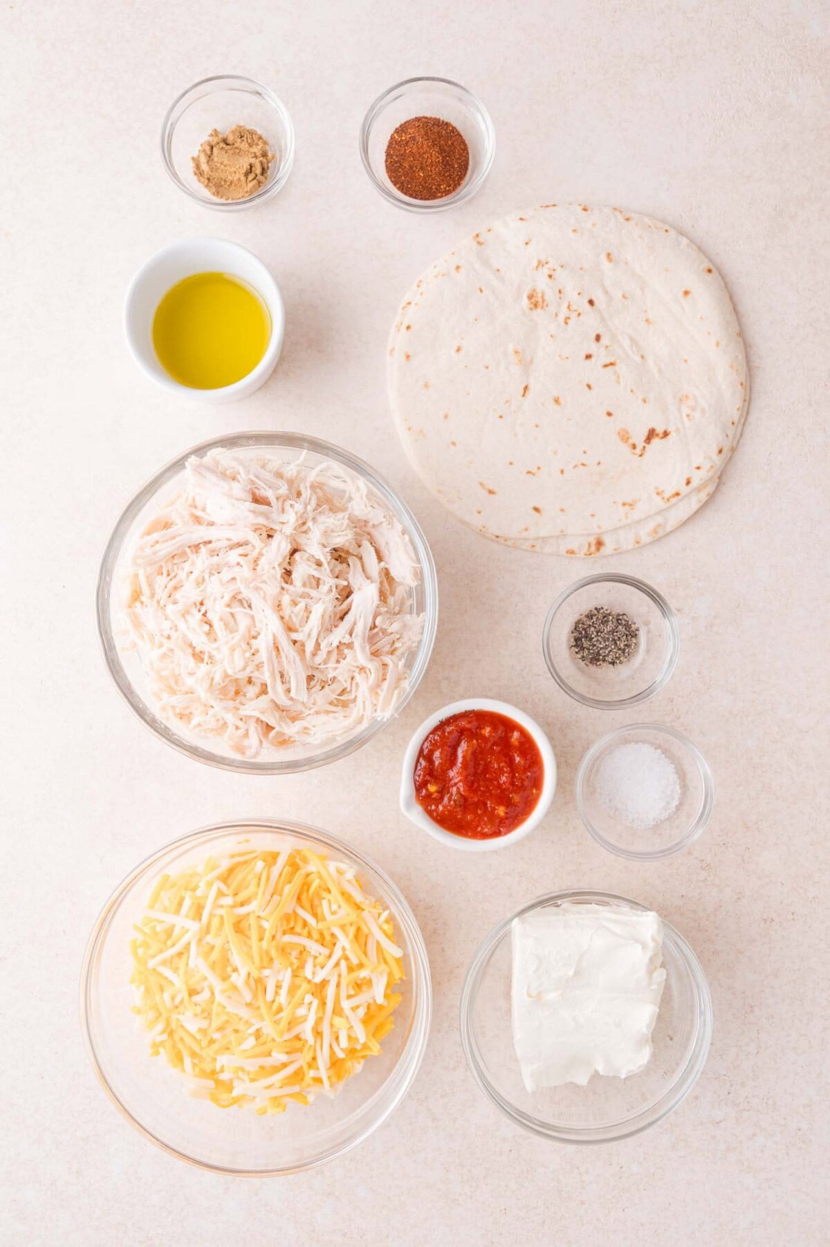Ingredients for a baked taquitos arranged on a white surface, including shredded chicken, shredded cheese, cream cheese, salsa, tortilla, spices, olive oil, salt, and pepper.
