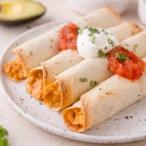 Four baked taquitos filled with a creamy mixture, topped with salsa, sour cream, and chopped herbs, served on a white plate with a sliced avocado in the background.