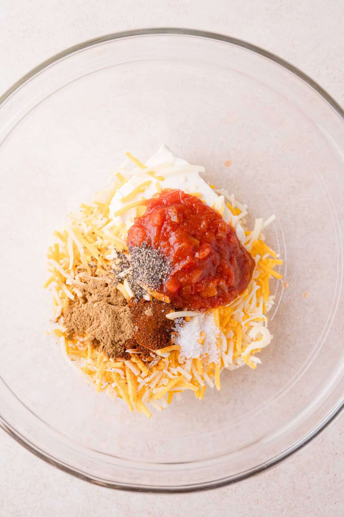 Clear glass bowl with shredded cheese, salsa, mayonnaise, and various spices on a light surface, ready to be mixed.
