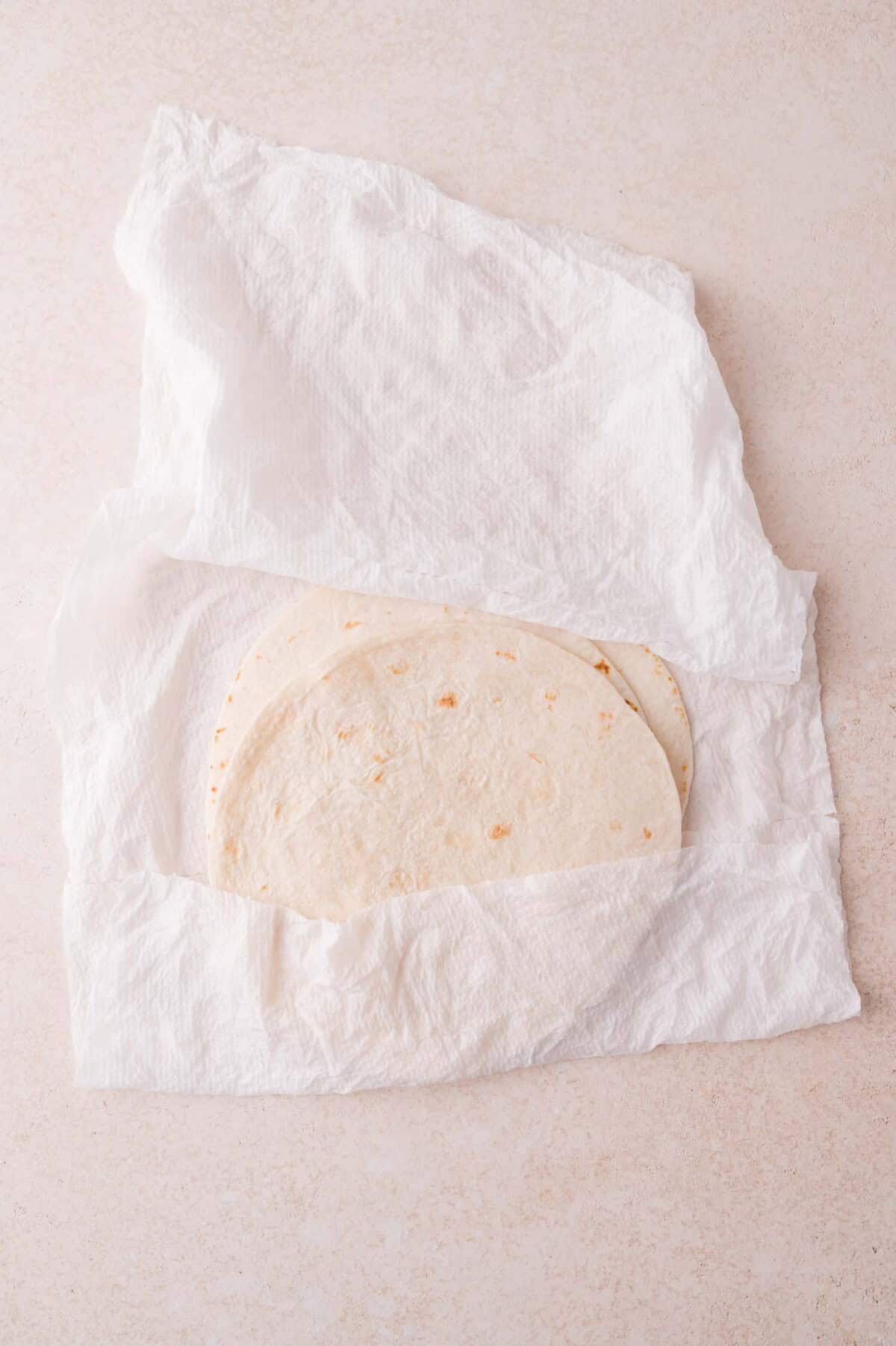 Two flour tortillas partially wrapped in a white paper towel, placed on a light-colored surface.
