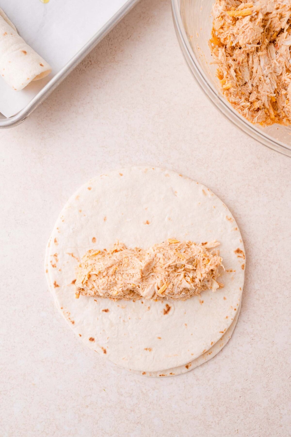 Flour tortilla with a portion of shredded chicken filling in the center, next to a bowl of chicken mixture and a baking sheet with a rolled tortilla.
