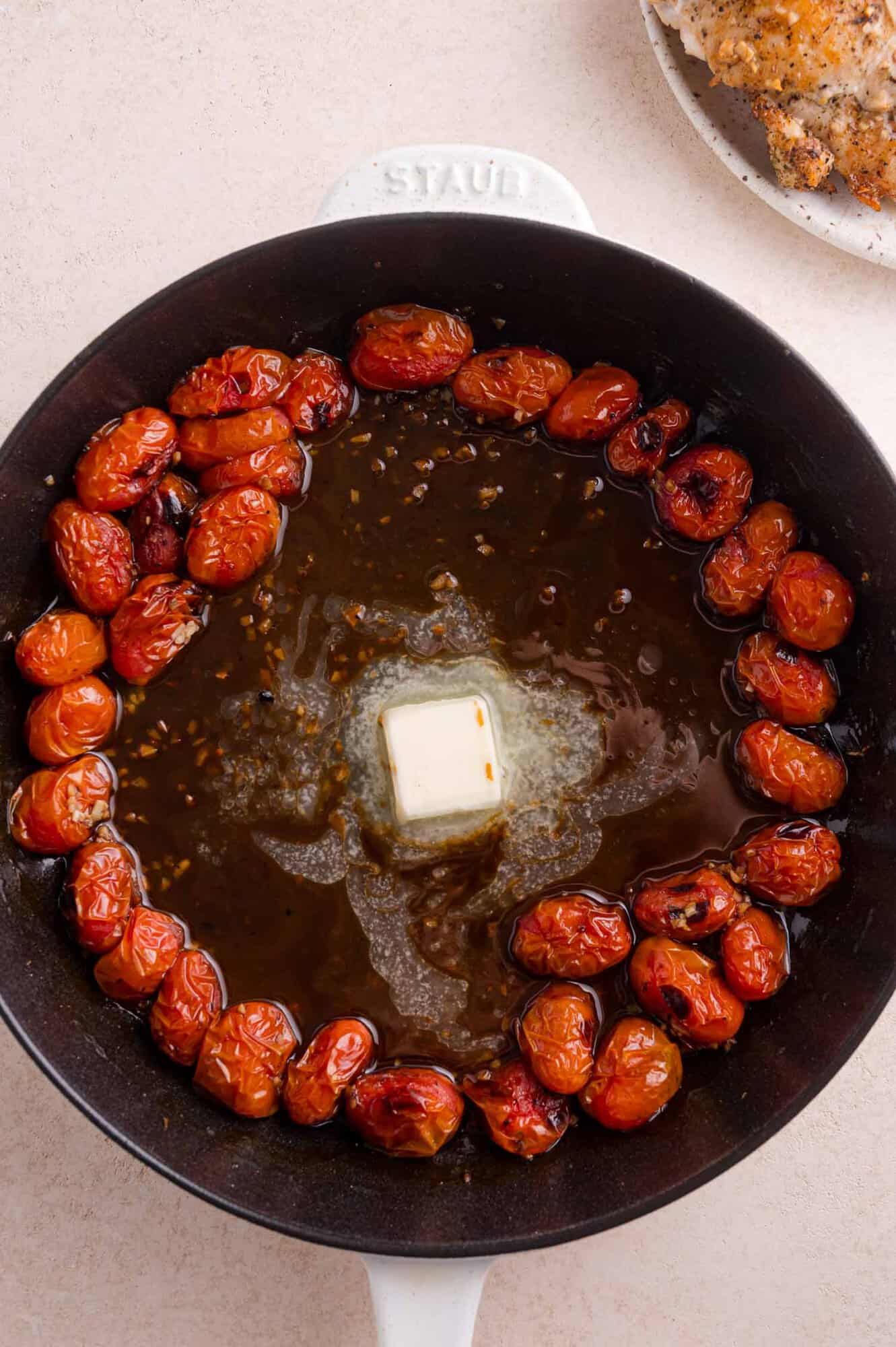 A cast iron pan with roasted cherry tomatoes, melted butter, and sauce in the center, on a beige surface.