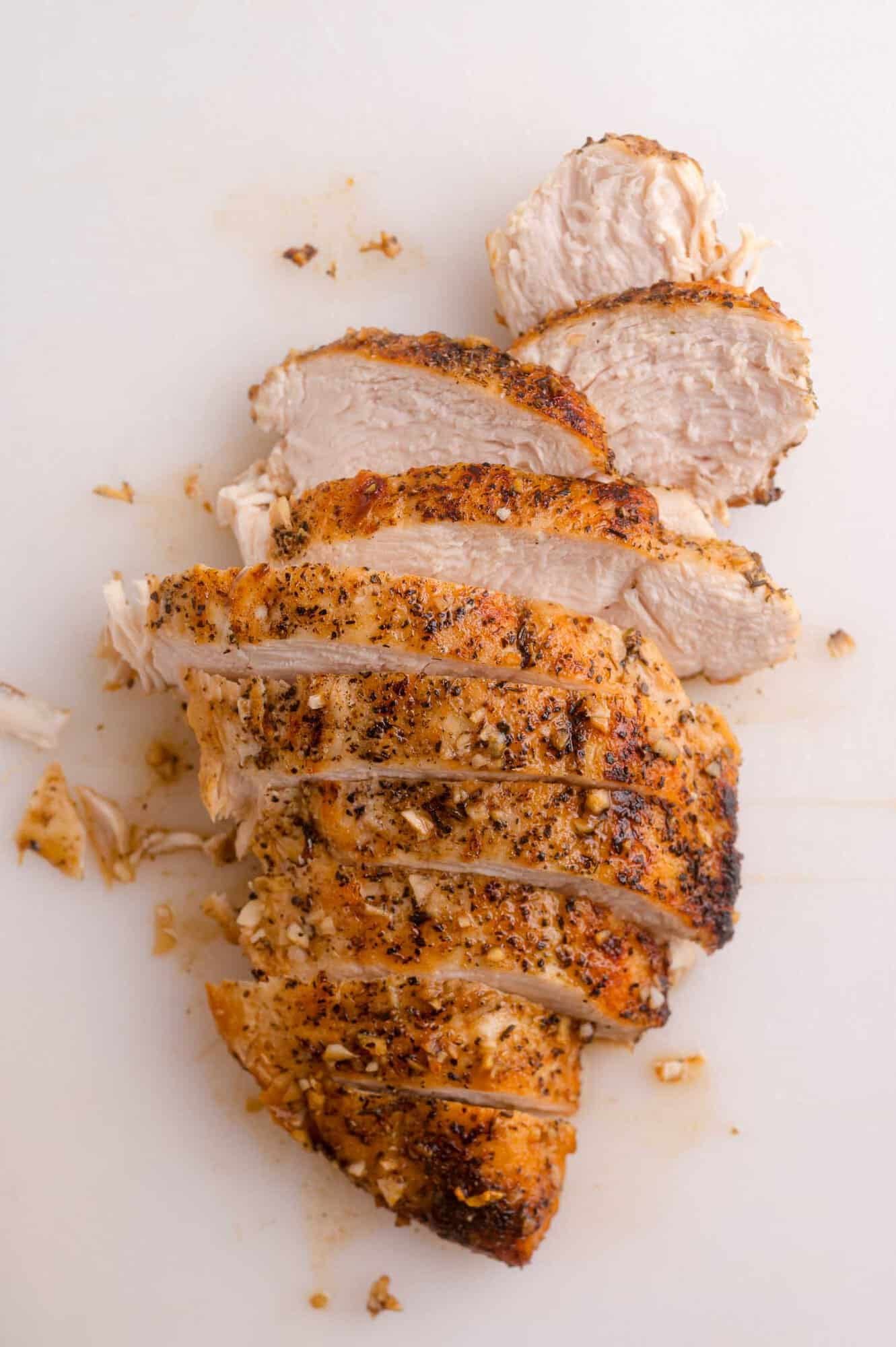 Sliced, cooked chicken breast seasoned with herbs and spices, arranged on a white surface.