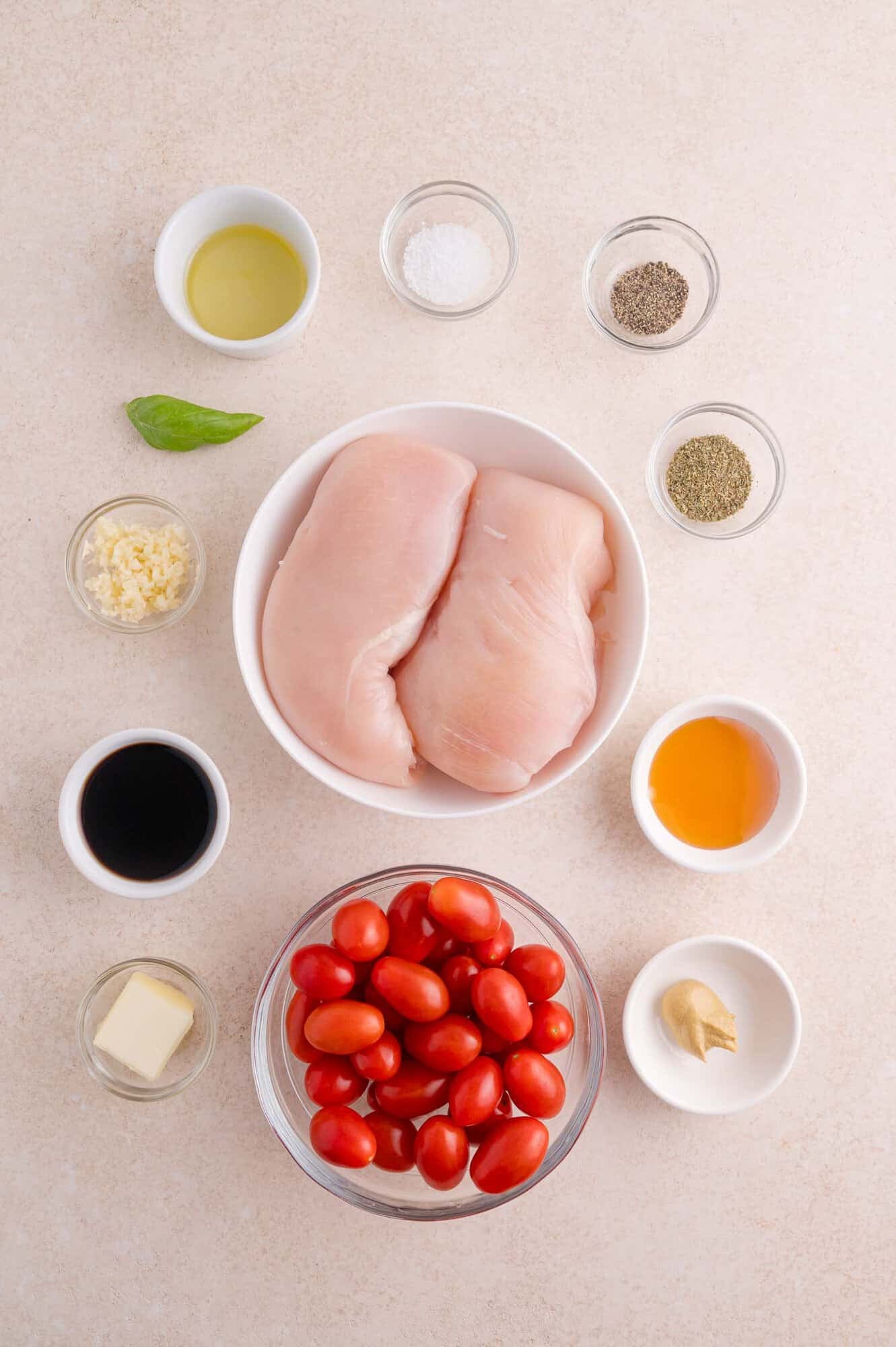 Two raw chicken breasts in a bowl, grape tomatoes in another bowl, and small dishes of oil, salt, pepper, garlic, vinegar, honey, butter, and mustard arranged on a light surface.