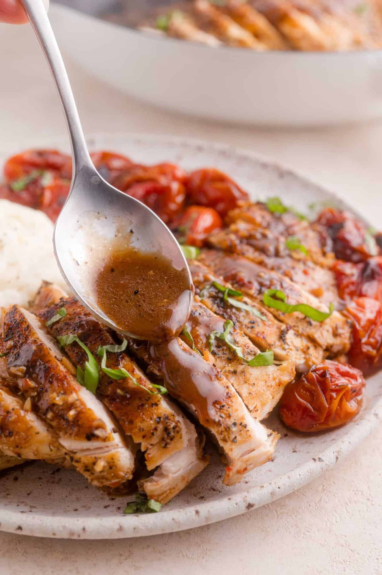 A spoon drizzles sauce over sliced grilled chicken breast, garnished with chopped herbs, with roasted tomatoes and a serving of mashed potatoes on the plate.