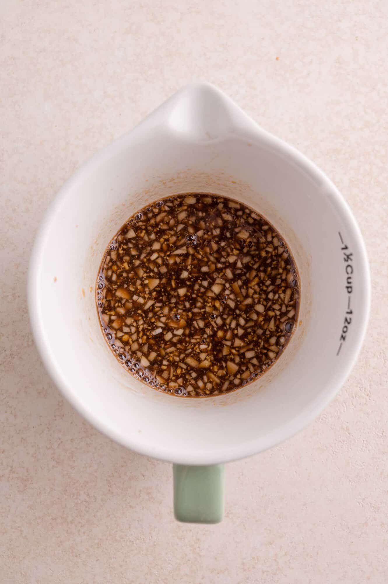 A white measuring cup containing a mixture of finely chopped garlic and balsamic-based sauce, viewed from above on a light surface.