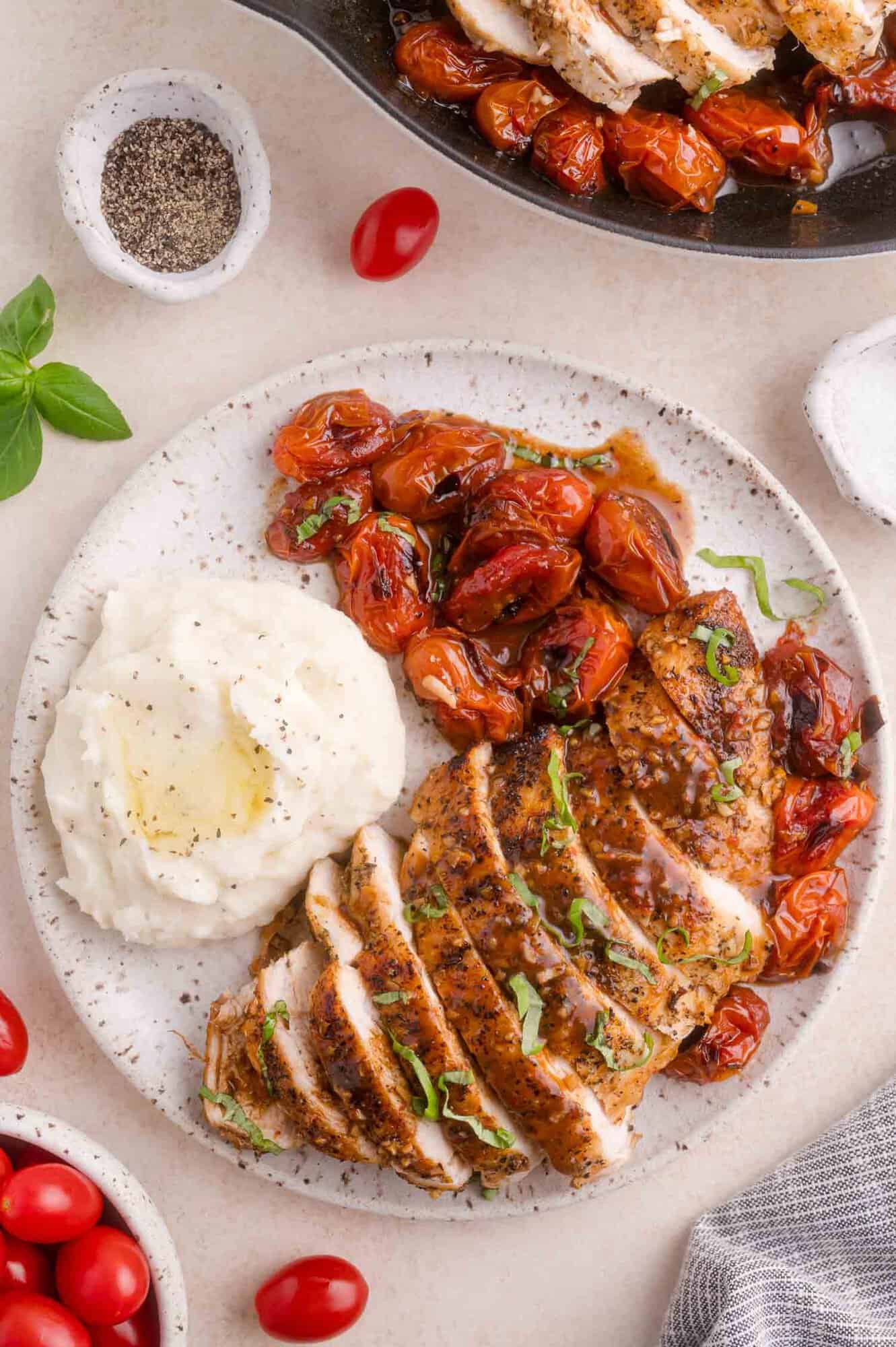 Sliced seasoned chicken breast with roasted cherry tomatoes and a serving of mashed potatoes on a white plate, garnished with fresh herbs.