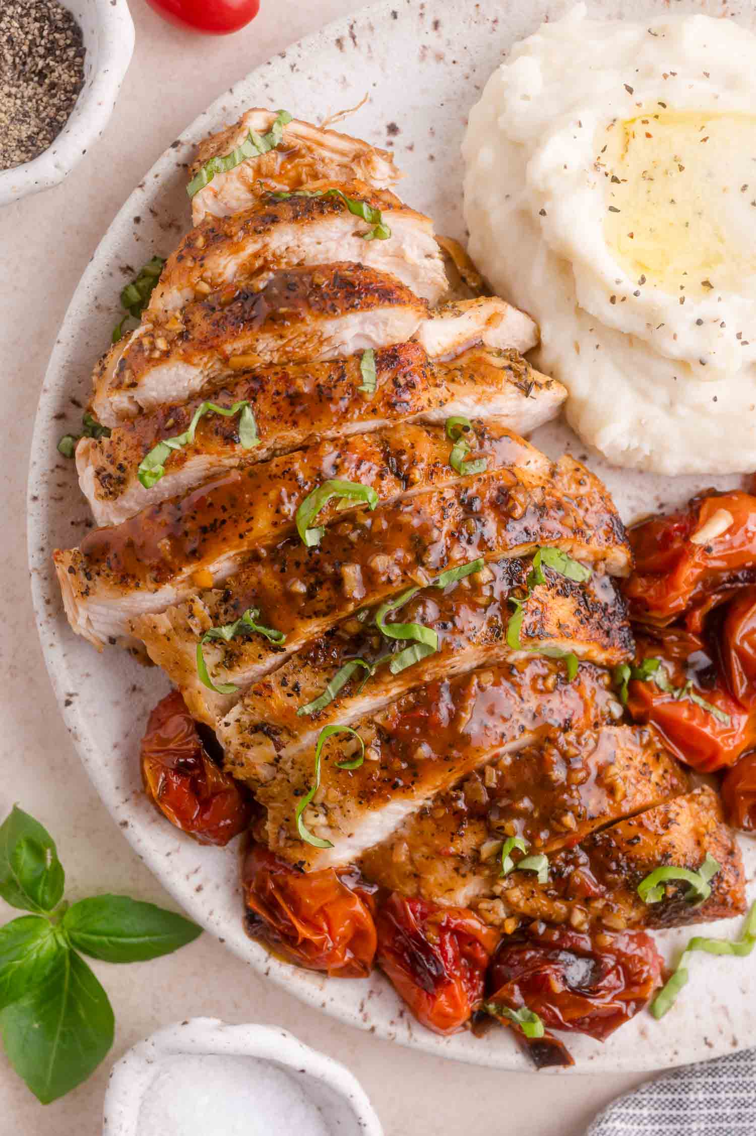 Sliced seasoned chicken breast with sauce, roasted tomatoes, and a serving of mashed potatoes on a white plate.