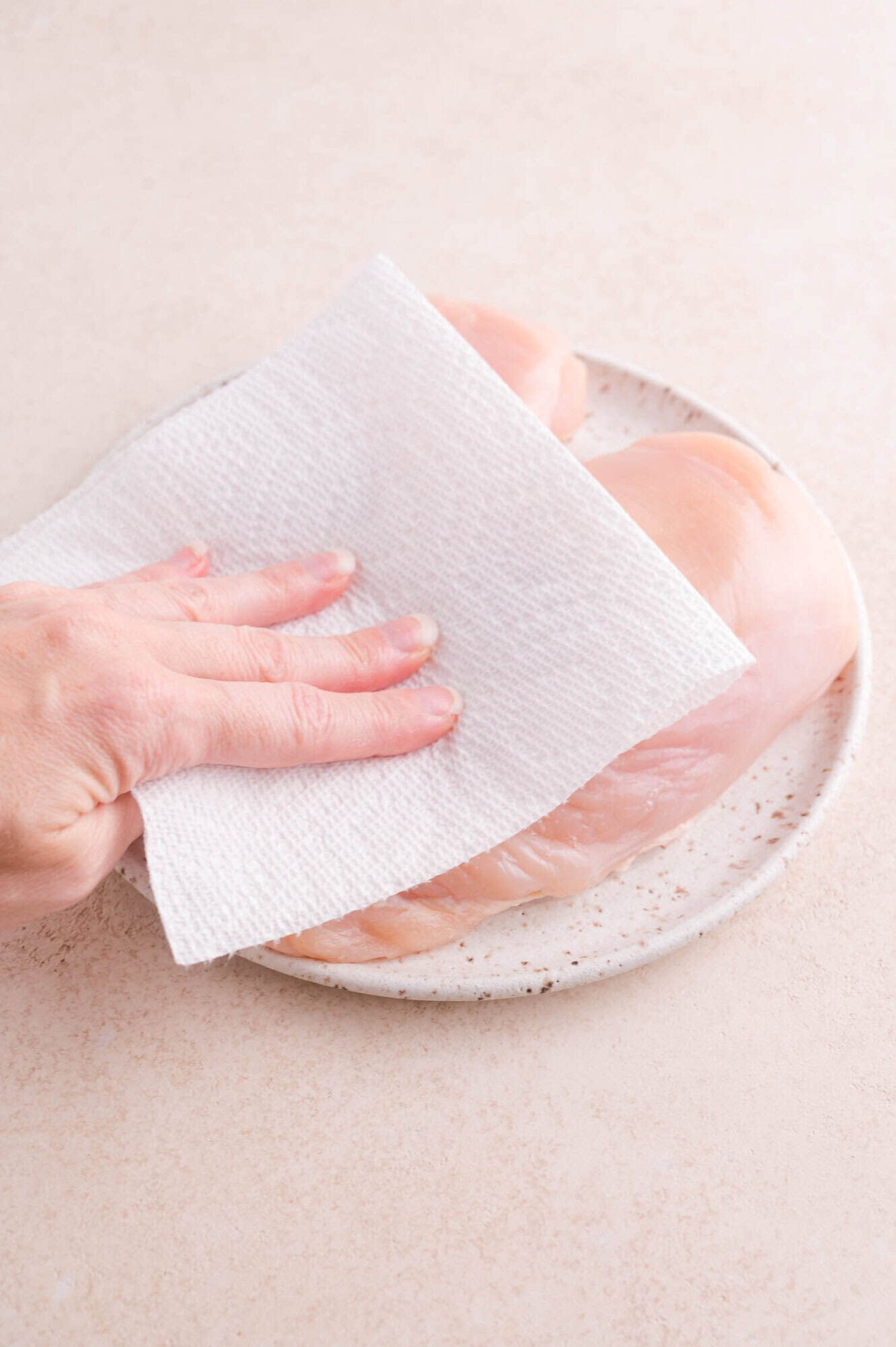 A hand uses a paper towel to pat raw chicken breasts dry on a speckled plate.