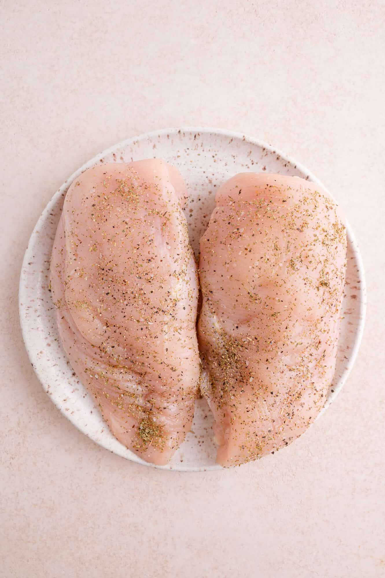 Two raw chicken breasts seasoned with pepper and herbs on a white plate, placed on a light-colored surface.