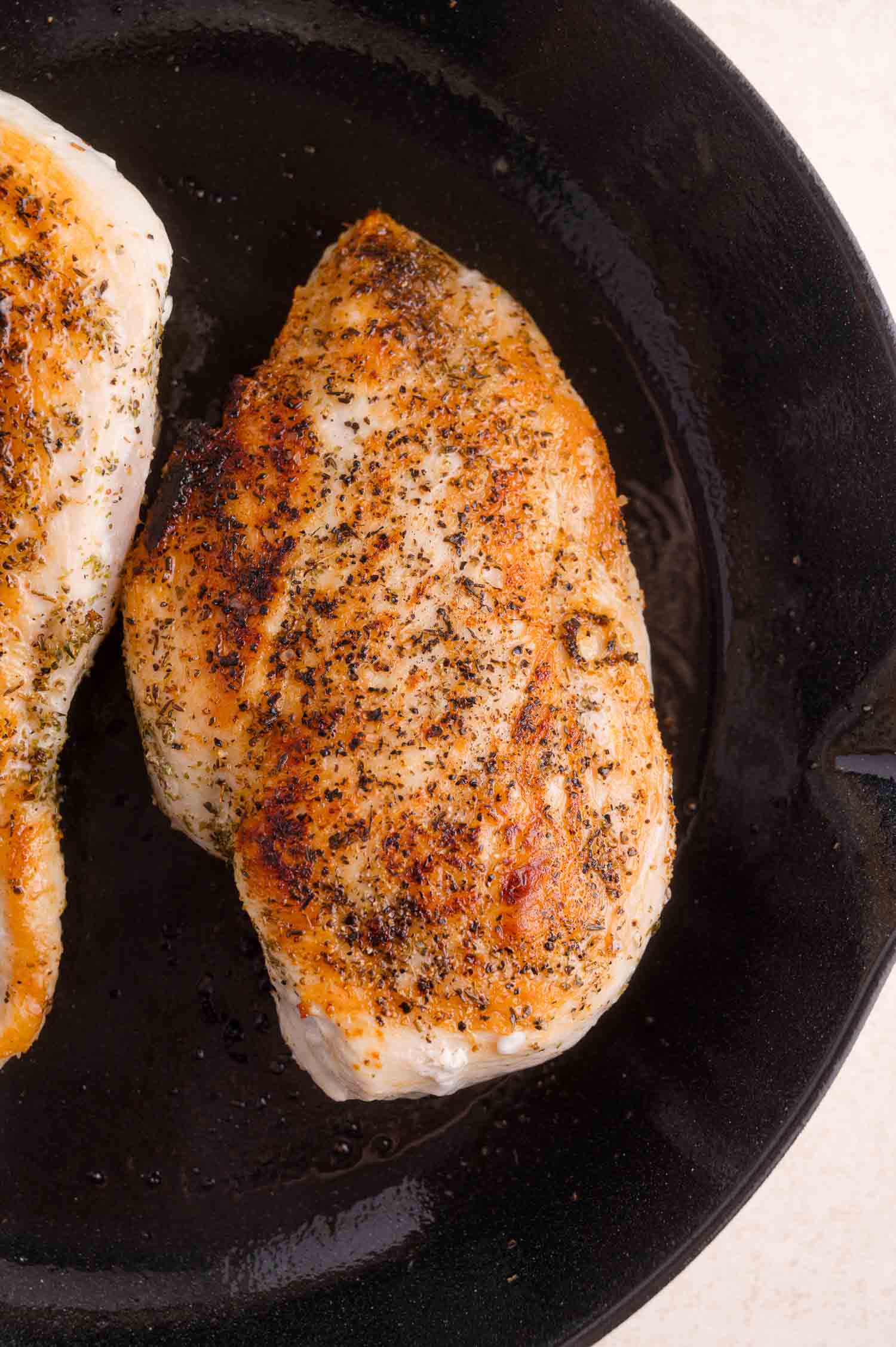 A seasoned, cooked chicken breast sits in a black cast iron skillet, showing a golden-brown, seared surface.