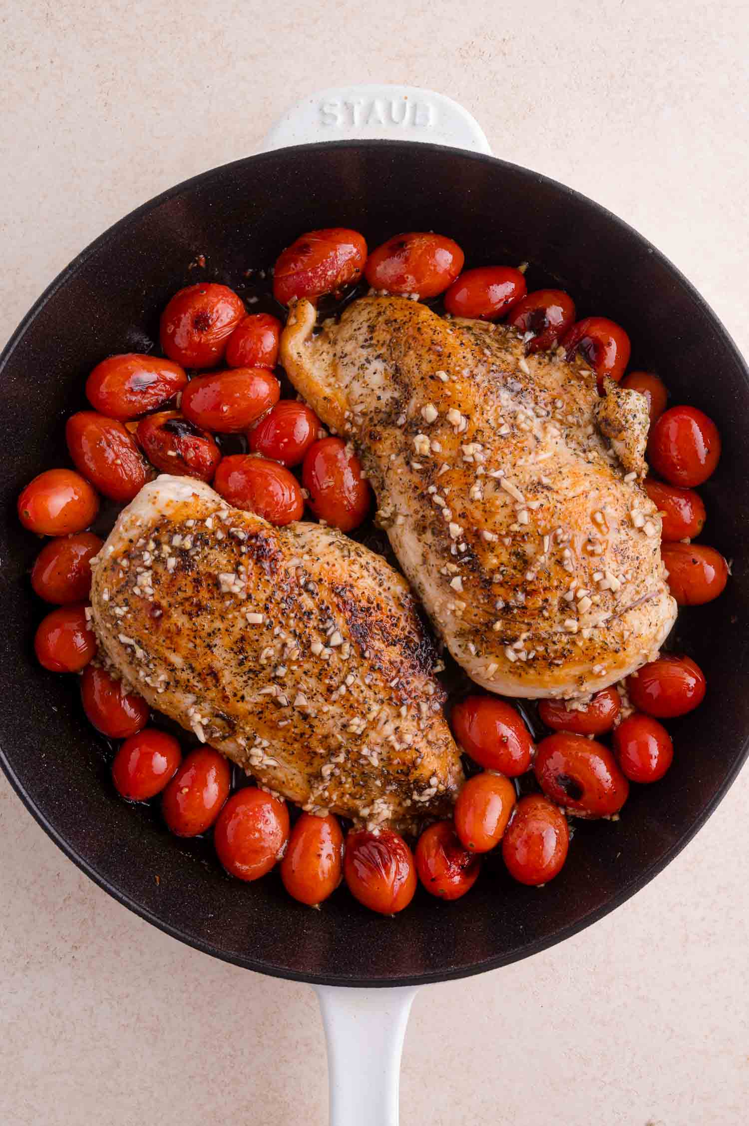 Two seasoned chicken breasts and roasted cherry tomatoes in a black skillet with a white handle.