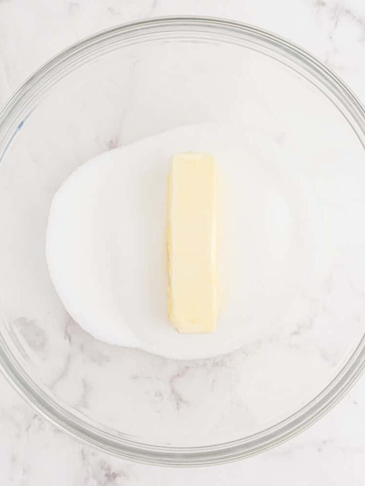 A glass bowl with a stick of butter on top of a pile of granulated sugar sits on a white marble surface next to a blue and white checkered towel.