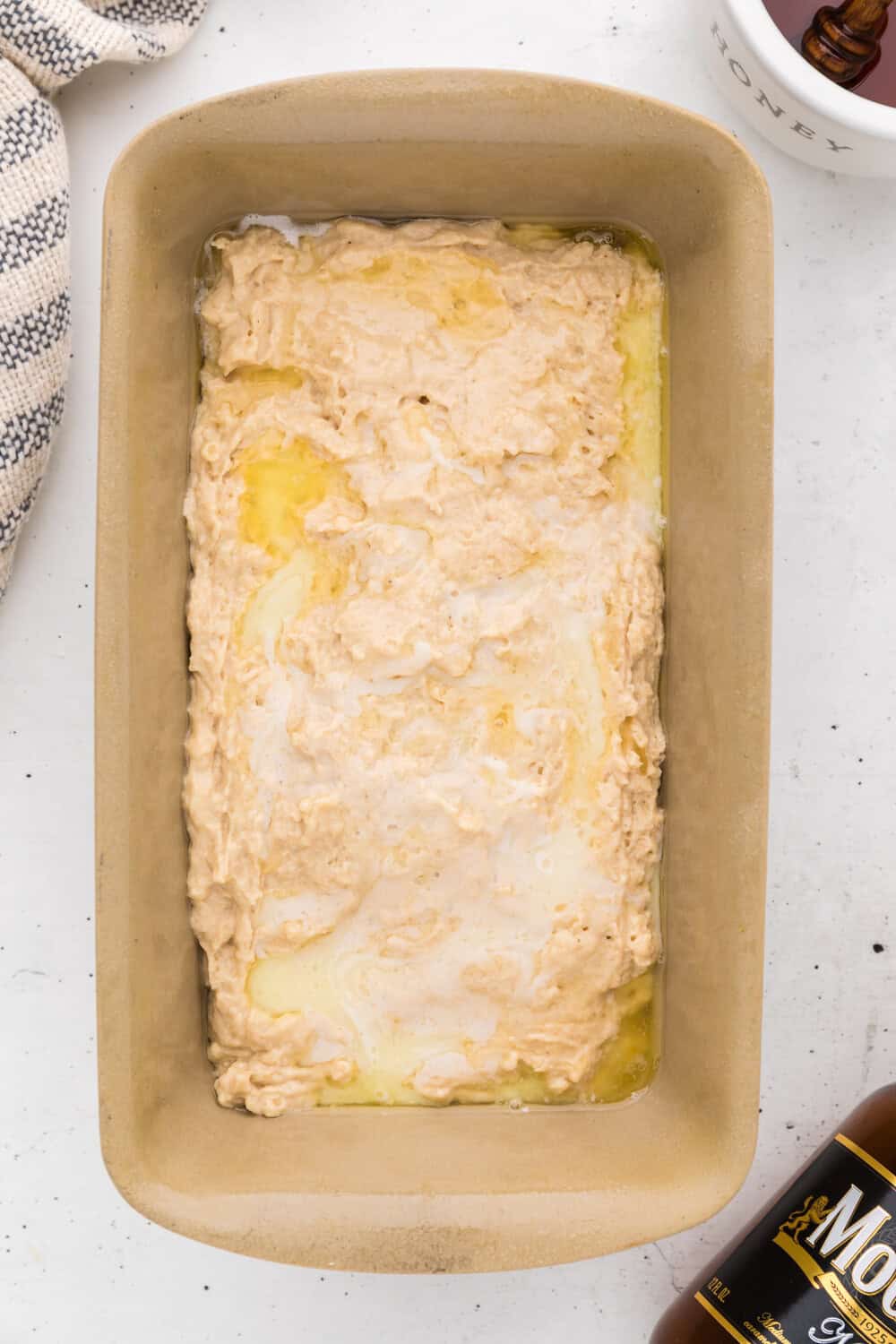Unbaked bread dough with swirls of melted butter in a rectangular loaf pan on a white surface, next to a towel and a bottle of honey.