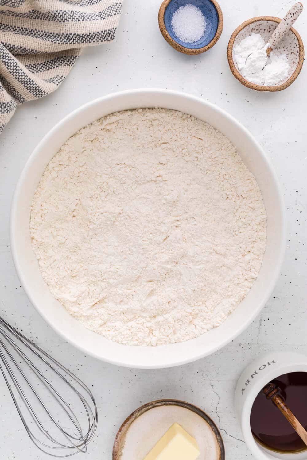 A large white bowl filled with flour is surrounded by a whisk, a dish of butter, a striped towel, salt, baking powder, and a mug of honey on a white surface.