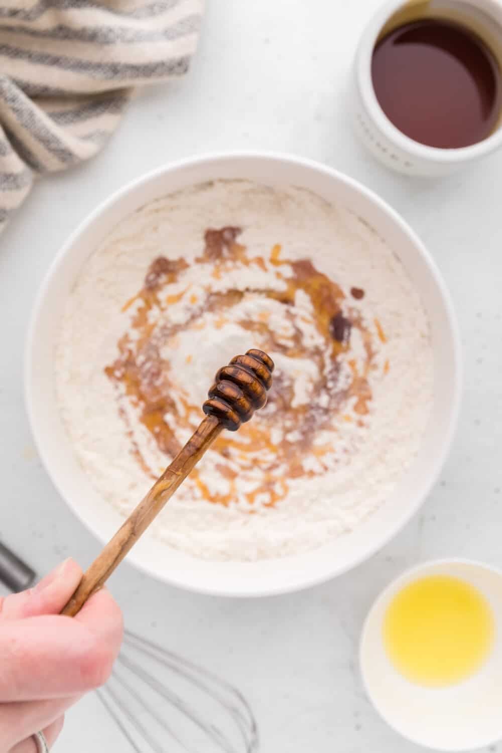 A hand holds a honey dipper over a bowl of flour with liquid ingredients, surrounded by bowls of oil and syrup on a white surface.