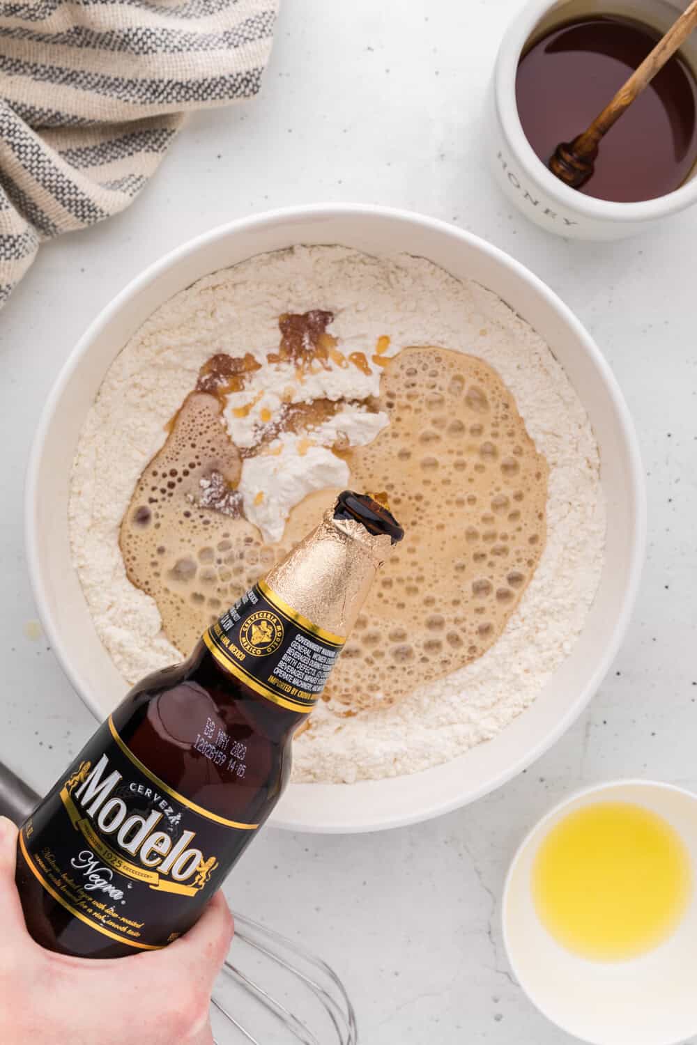 A hand pours Modelo Negra beer into a bowl of flour, honey, and other ingredients, with melted butter and a honey jar nearby on a white surface.