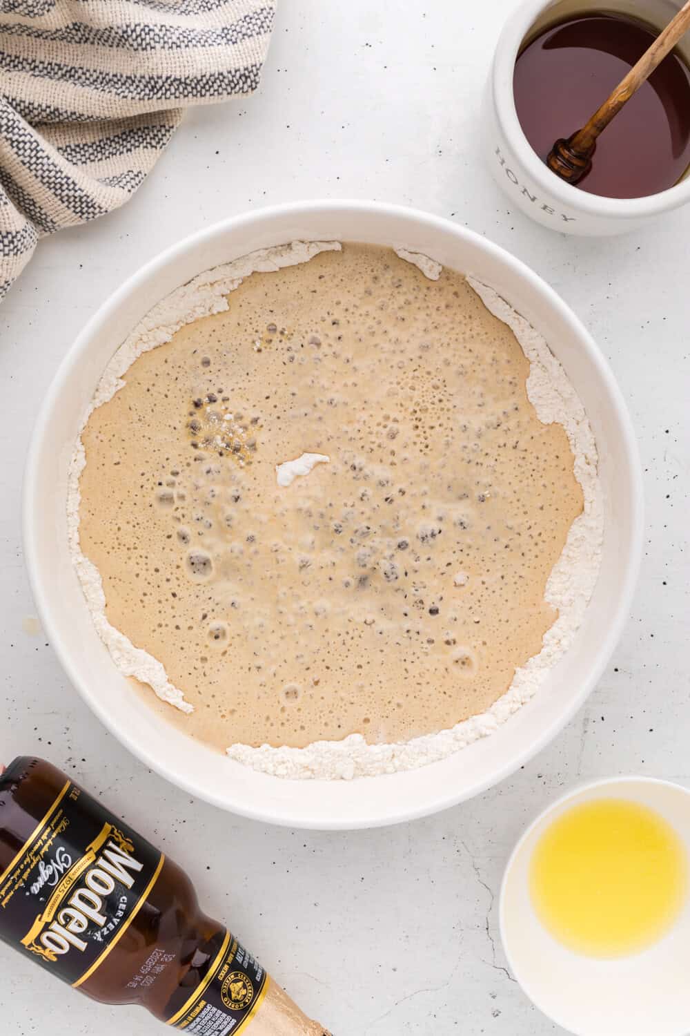 A mixing bowl with flour, beer, and bubbles sits on a countertop beside a bottle of Modelo, a small bowl of melted butter, and a jar of honey with a dipper.