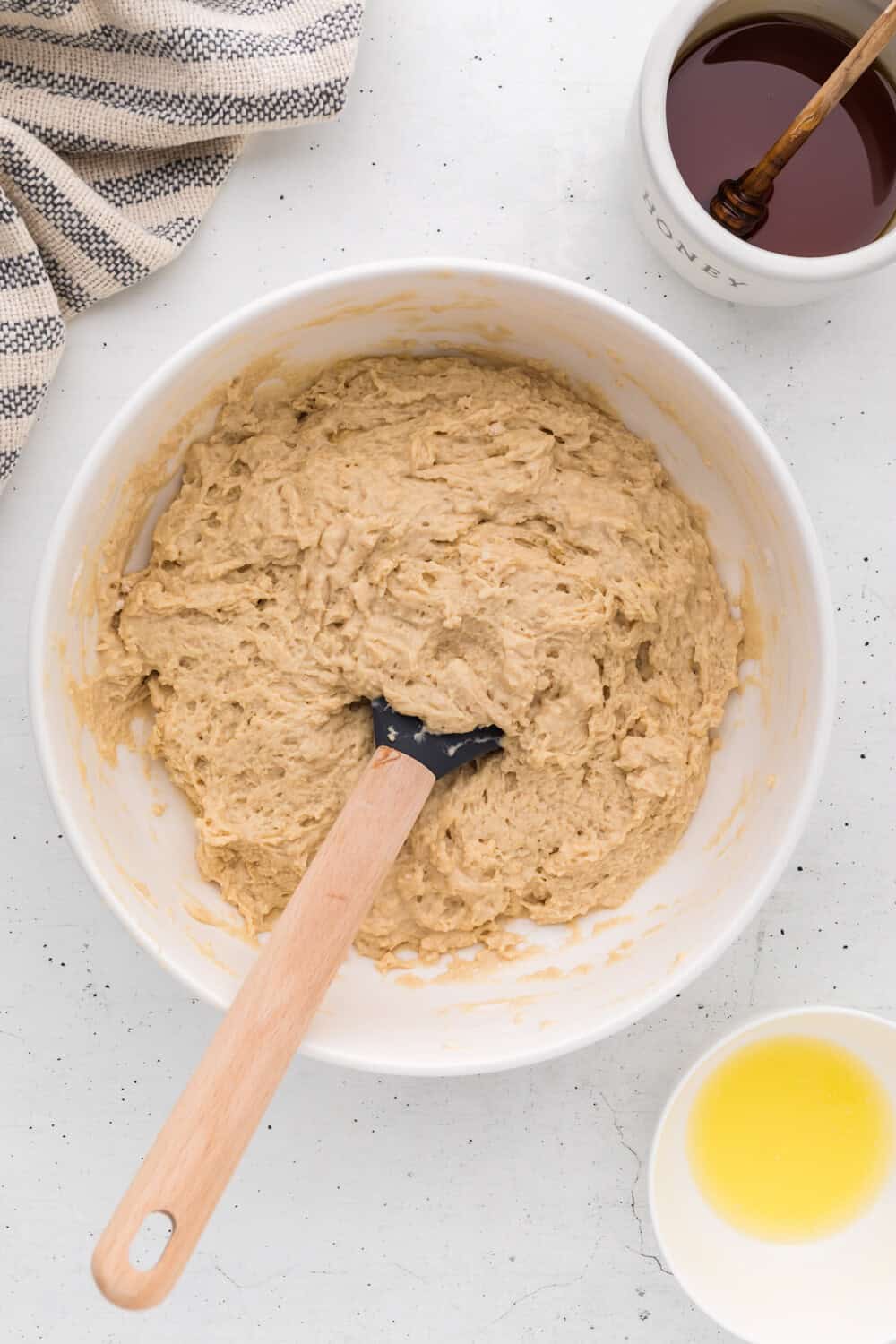 A mixing bowl with dough and a spatula, next to a jar of honey with a dipper, a small bowl of melted butter, and a striped towel.