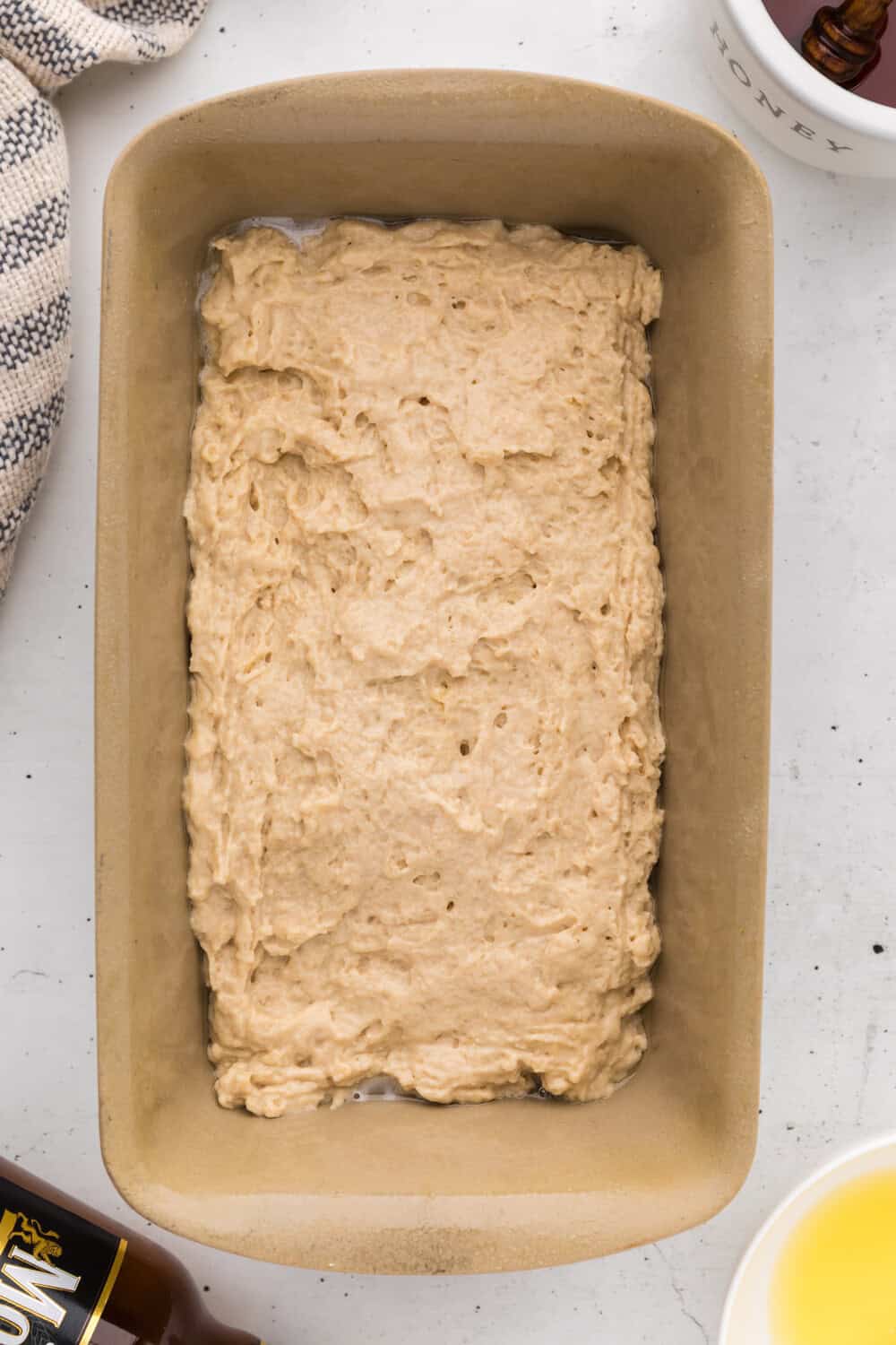 Unbaked bread dough spread evenly in a rectangular loaf pan, surrounded by a towel, a cup, and a bottle on a white surface.
