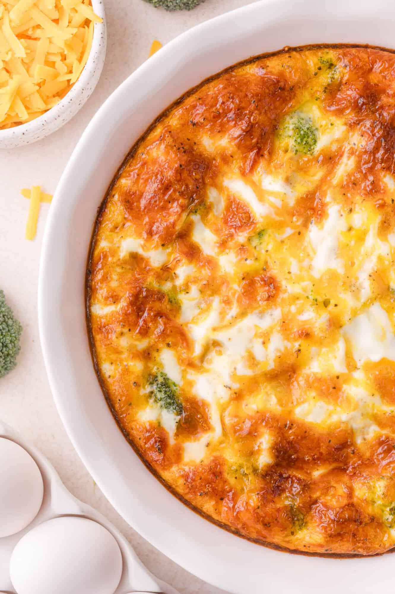 Baked broccoli and cheese casserole in a white dish, with cheddar cheese, raw broccoli, and eggs nearby on a light surface.