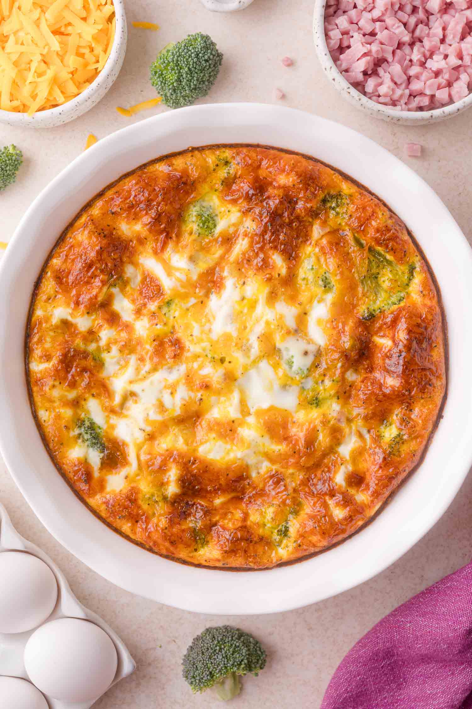 A baked crustless quiche with a golden-brown top, visible broccoli pieces, and surrounded by bowls of shredded cheese, diced ham, eggs, and broccoli florets on a light surface.
