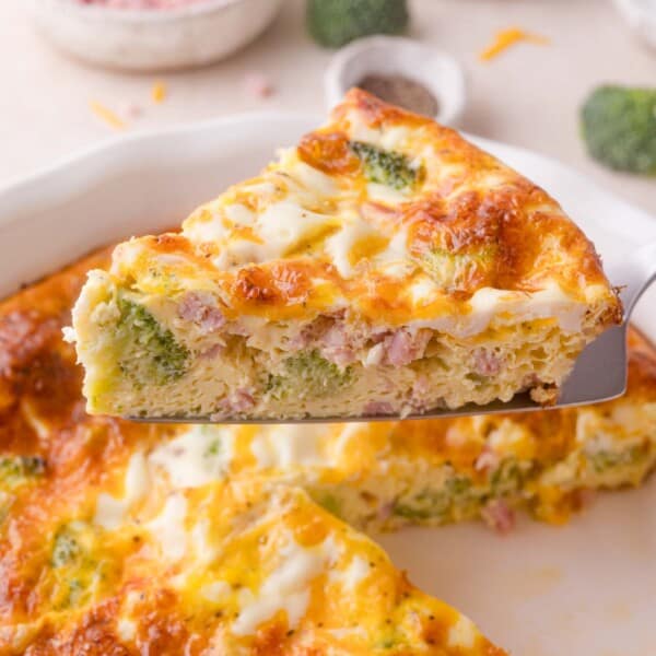 A close-up of a slice of broccoli, ham, and cheese crustless quiche being lifted from a pie dish.