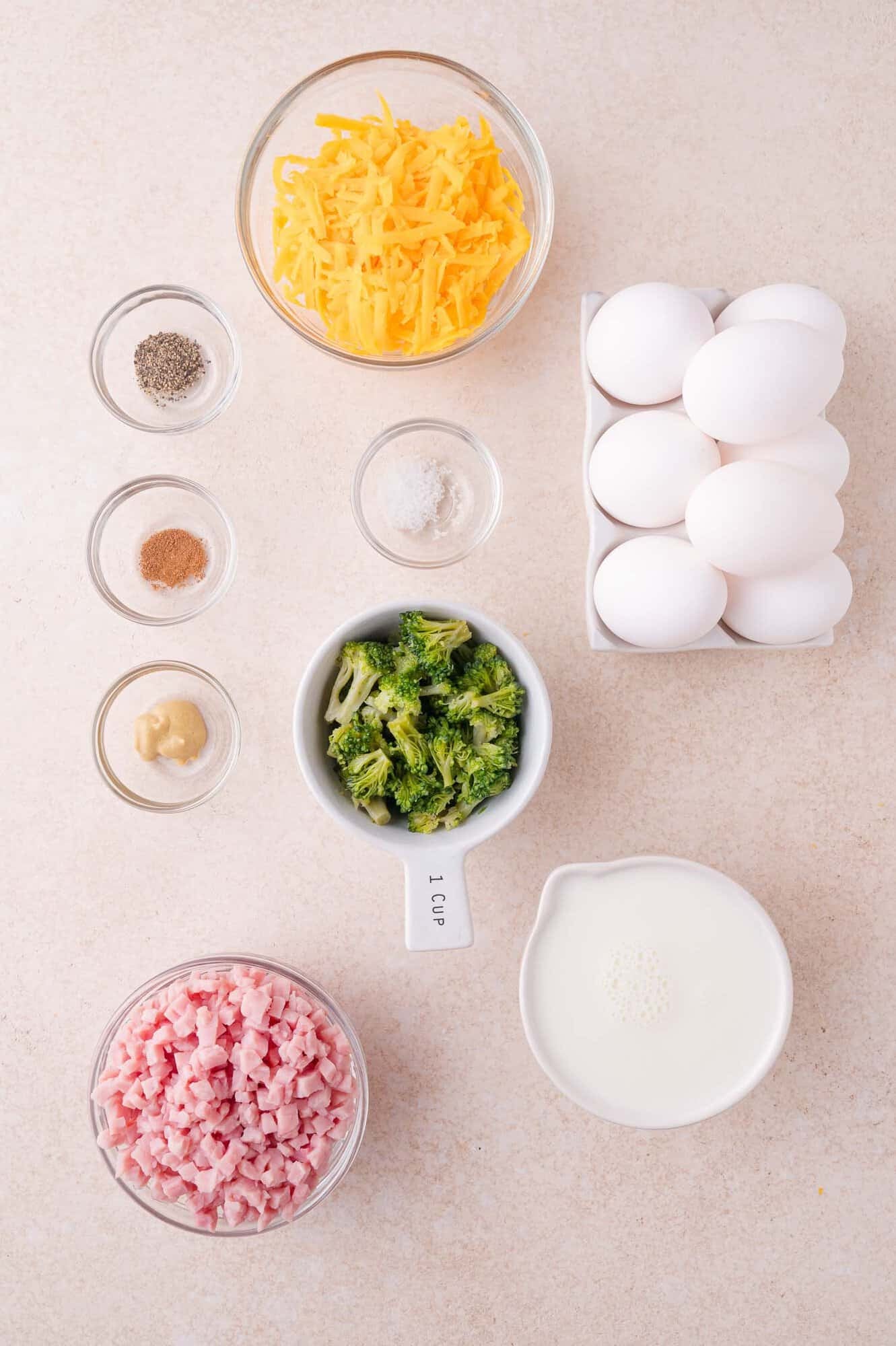 Top-down view of ingredients: shredded cheese, eggs, broccoli, diced ham, milk, mustard, pepper, nutmeg, and salt arranged on a light surface.